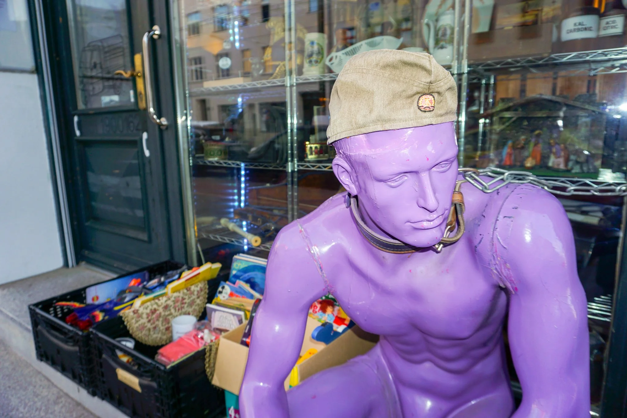 Eine schunkelnde Statue in lila Farbe, die einen beige Hut trägt, in einer Straßenszene bei Tag, umgeben von einem Schaufenster mit Gegenständen und Spielzeug. Im Hintergrund sind Gebäude sichtbar.