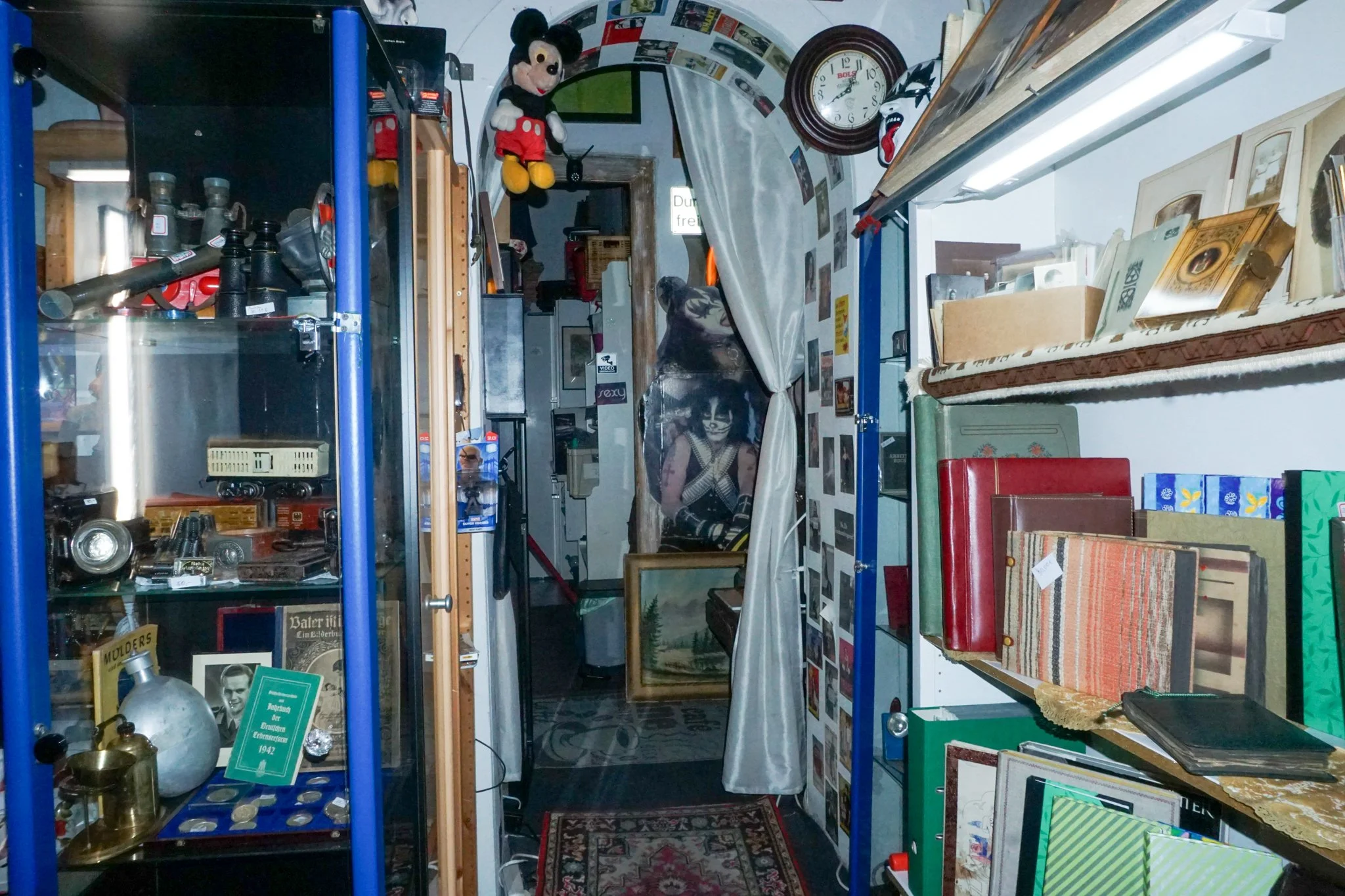 Ein Raum mit alten Büchern, dekorativen Gegenständen, einem Wandbild und Puppen, darunter Mickey Mouse und ein Masken-Illusionsseil. Es sieht aus wie ein Antiquitäten- oder Flohmarktrraum.