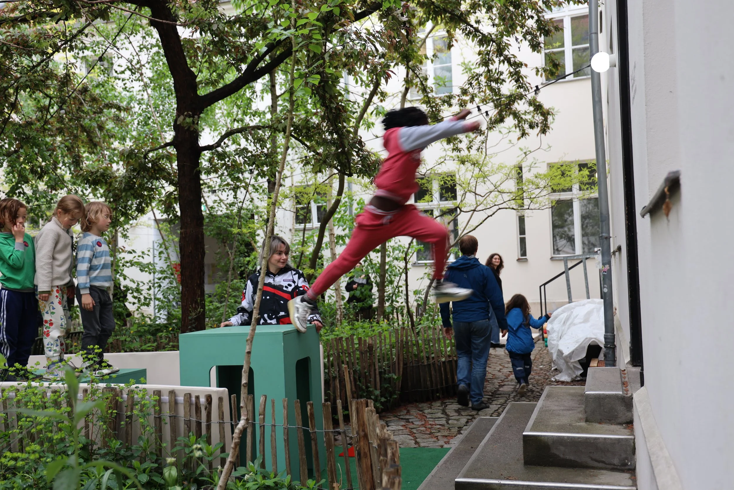 Junge beim Klettern an einem Seil in einem Garten mit mehreren Erwachsenen und Kindern, Bäumen und grünem Umfeld.