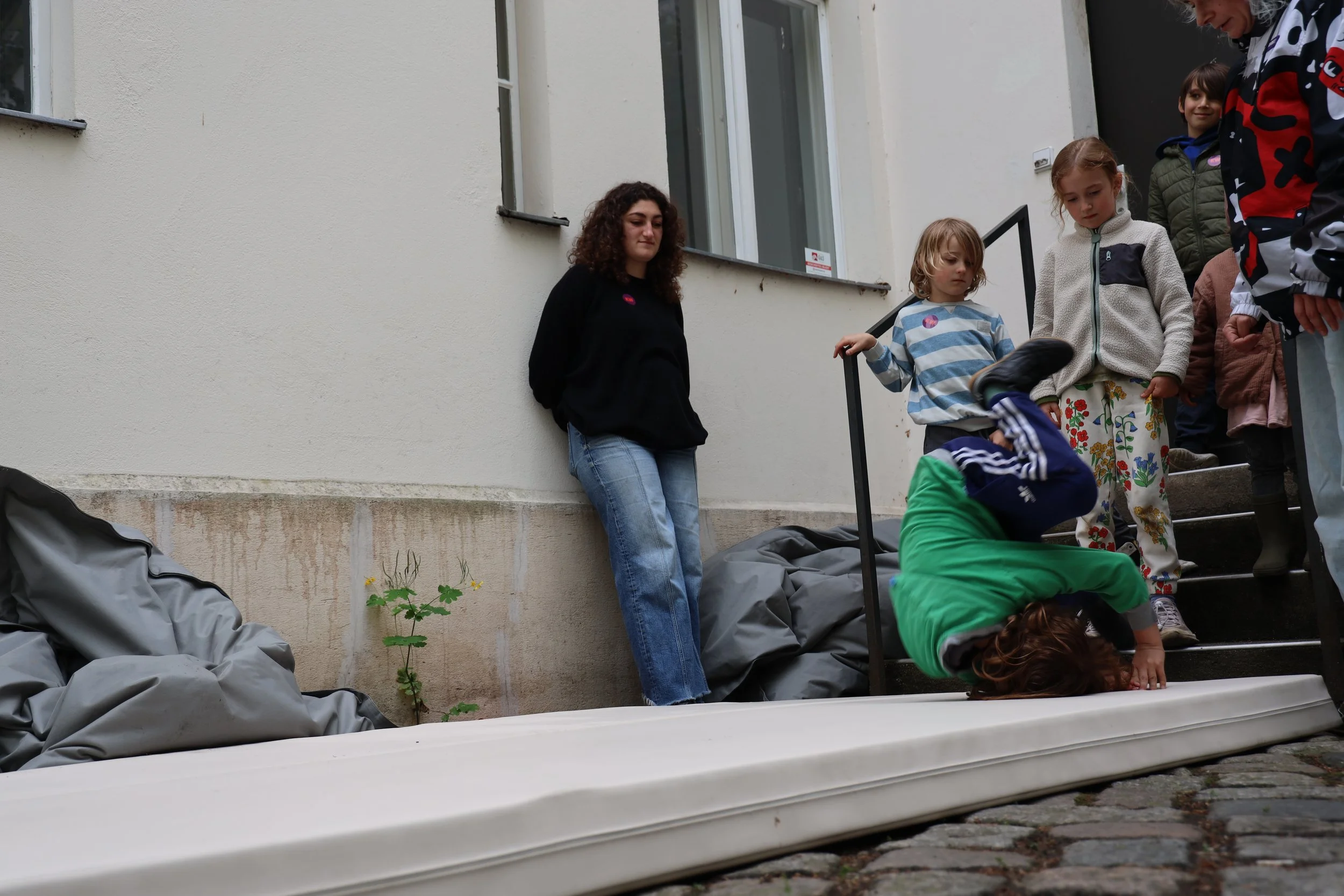 Kinder und Erwachsene auf einer Treppe, einige schauen nach unten, andere unter einem failierten Versuch, sich umzudrehen, liegen auf dem Boden, mit schwarzen Säcken und einem grünen Kleidungsstück.