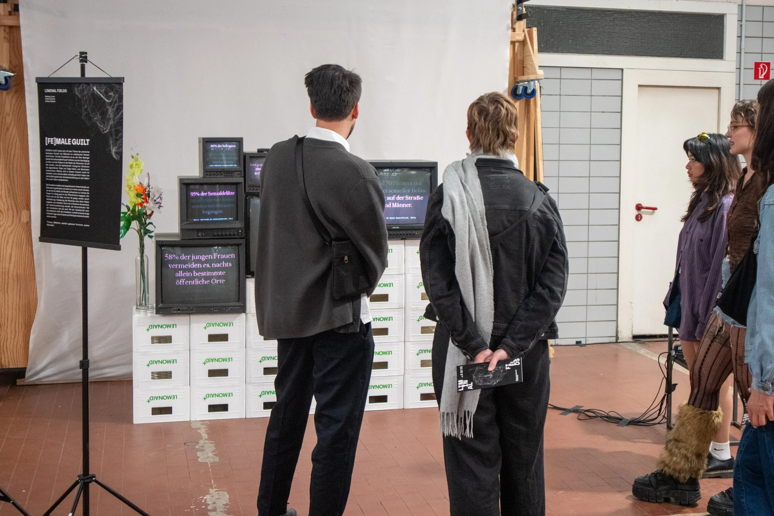 Menschen stehen in einer Ausstellung vor einem Bildschirm, der Statistiken zeigt. Es gibt eine Garderobe und einen schwarzen Poster mit Informationen auf der linken Seite.