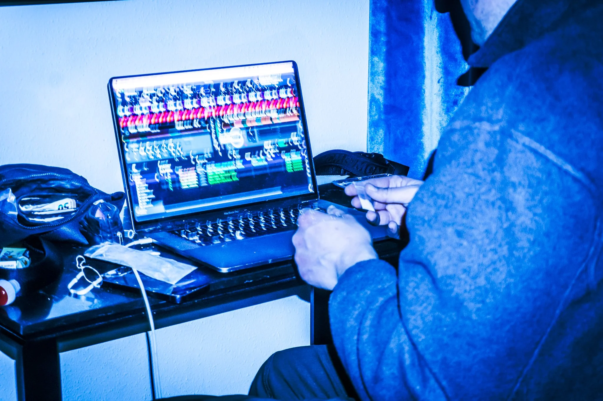 Eine Person arbeitet an einem Laptop mit einem hell leuchtenden Bildschirm auf einem Tisch, umgeben von verschiedenen Gegenständen. Der Raum ist in blauen Tönen beleuchtet.