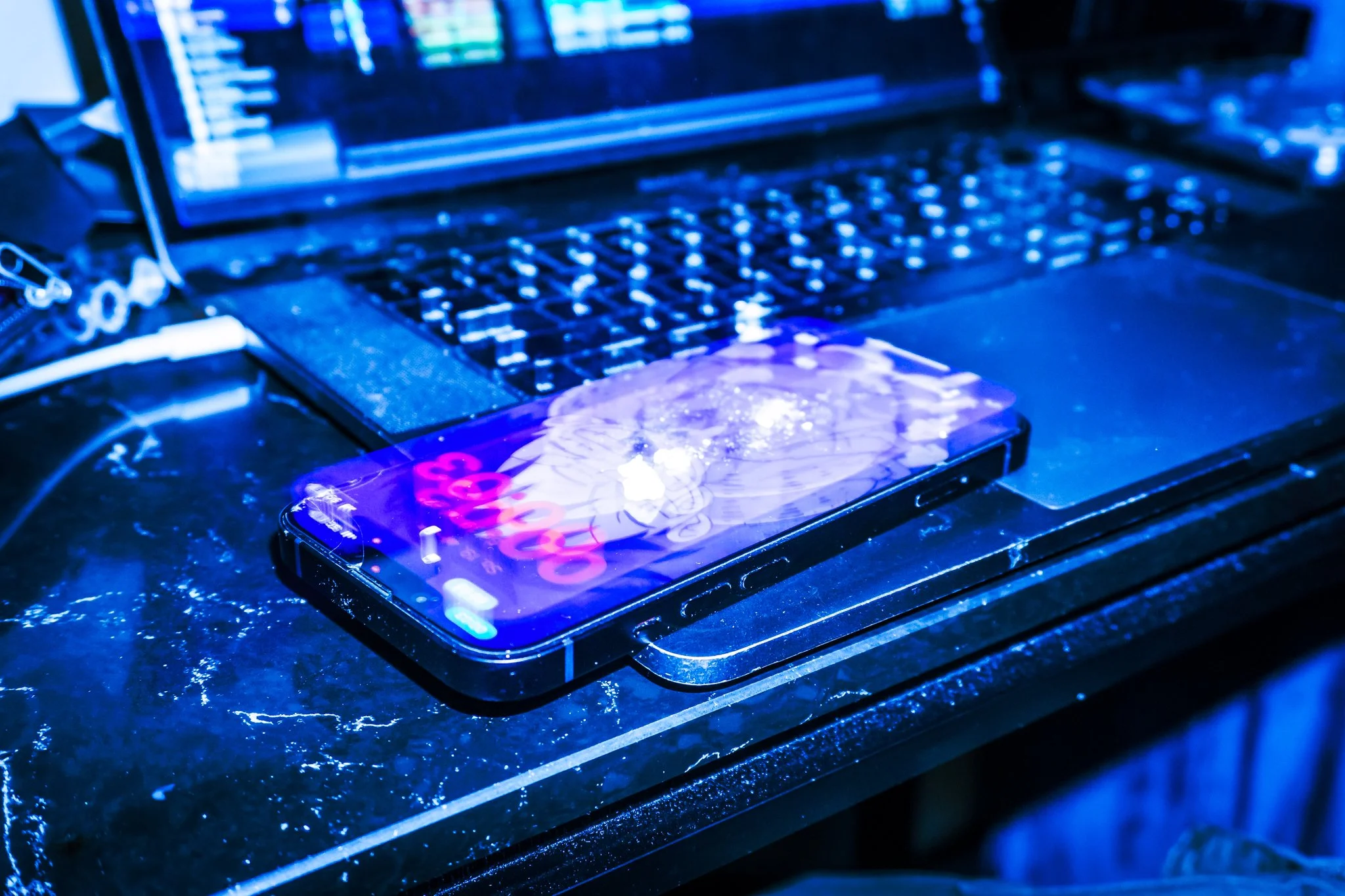 Ein Smartphone liegt auf einem dunklen, nassen Tisch. Der Bildschirm zeigt ein leuchtendes, grafisches Display, während im Hintergrund eine Laptop-Tastatur zu erkennen ist, die von blauem Licht beleuchtet wird.