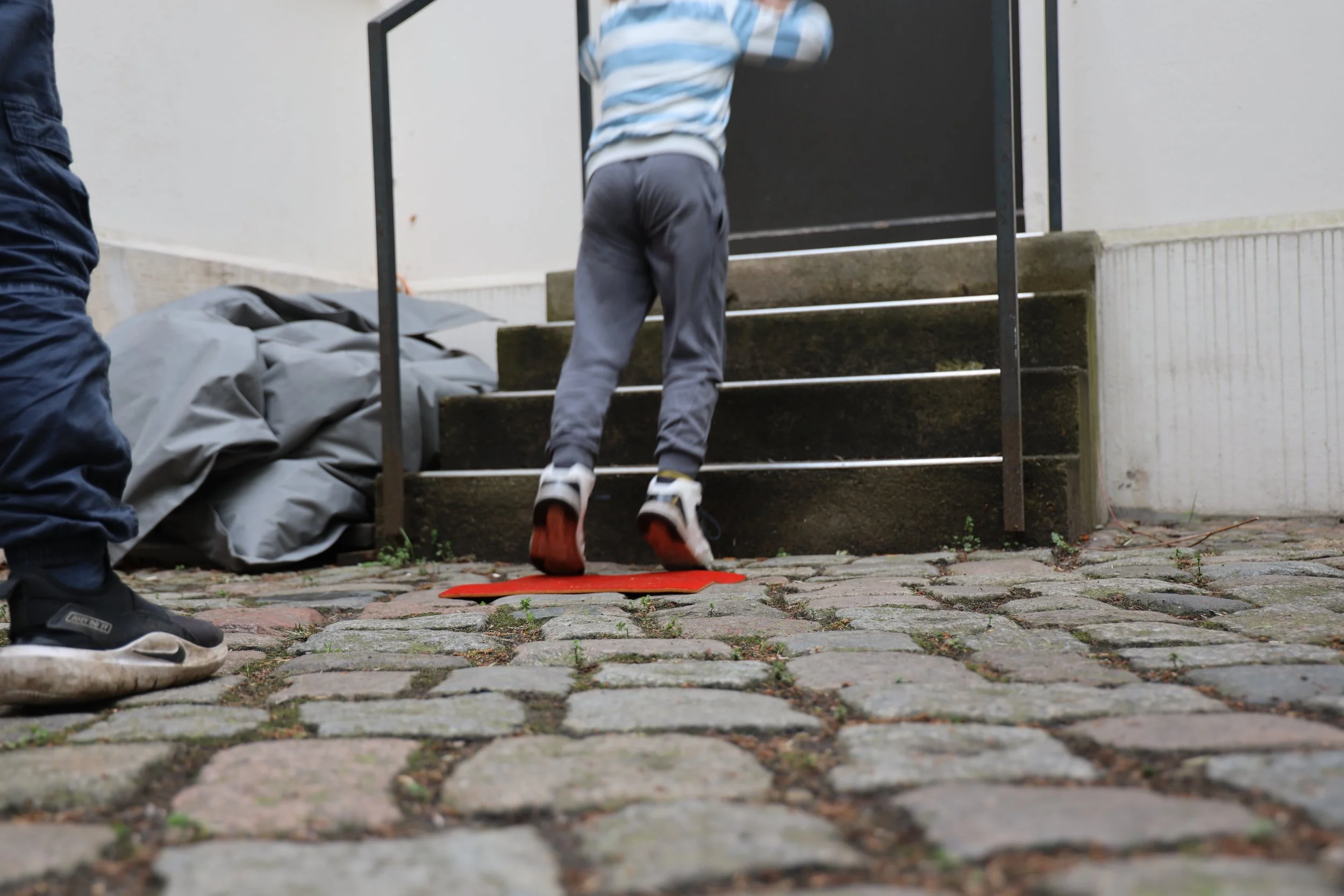 Kinder klettern auf eine Treppe aus Holz mit schwarzen Metallgeländern, während andere auf dem Boden stehen, darunter ein rotes Kissen. Der Bereich ist im Freien mit einer Pflastersteinfläche. Es gibt eine graue Abdeckung oder einen Rucksack auf dem 
