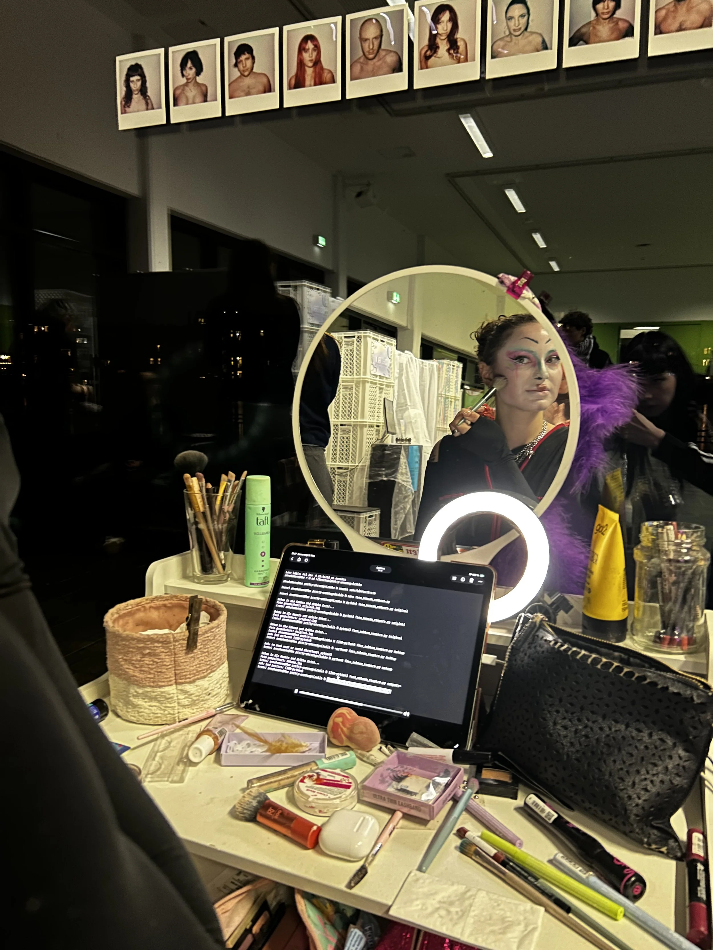 Ein Spiegel zeigt eine Frau mit künstlerischem Make-up und Kostüm, umgeben von Make-up- und Kosmetikartikeln auf einem Tisch, im Hintergrund sind mehrere Menschen und Fotoporträts an der Wand.