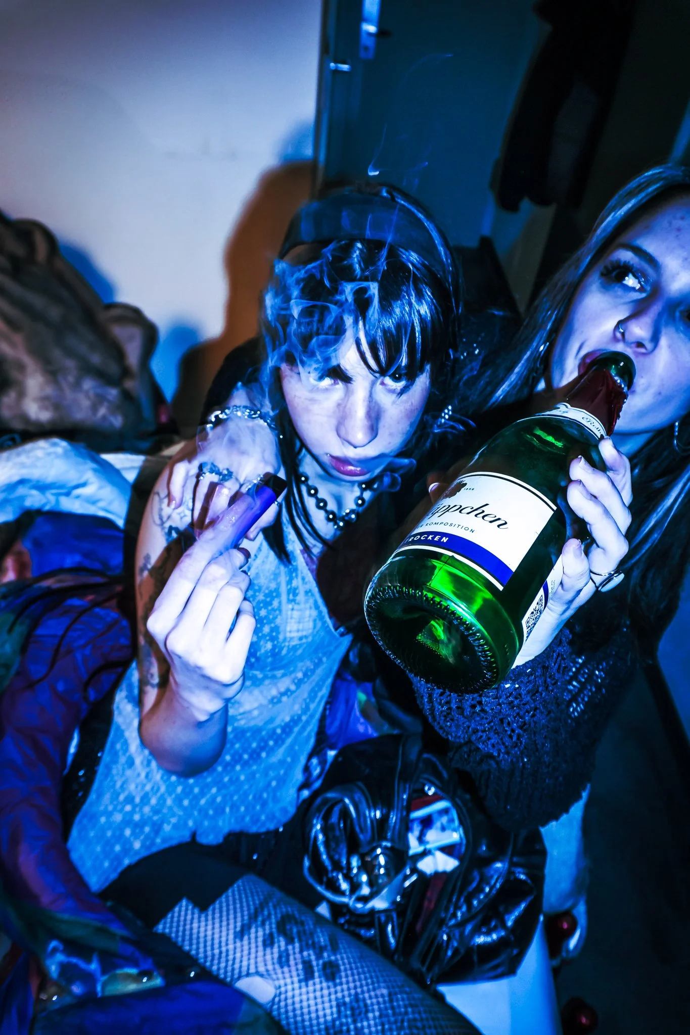 Zwei junge Frauen in einer Party oder Club, eine trinkt Alkohol aus einer Flasche, die andere raucht eine Zigarette. Sie sind in einem dunklen Raum mit Blaulicht