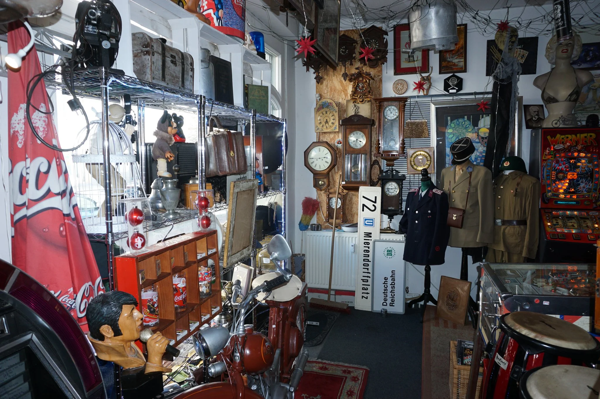 Ein Raum voller verschiedenster Antiquitäten, Sammlerstücke und Dekorationsgegenstände, darunter Wanduhren, Puppen, Motorräder, eine Figur mit Mikrofon, Spielautomaten, und andere Dekorationsartikel in einem Vintage- oder Flohmarktladen.