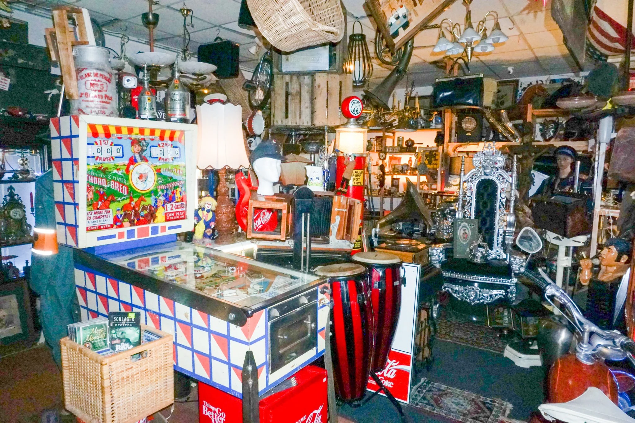 Verschiedene alte und antike Gegenstände und Dekorationen in einem Raum, inklusive Spielautomat, Lampen, Flaschen, Möbel und kleine Skulpturen.