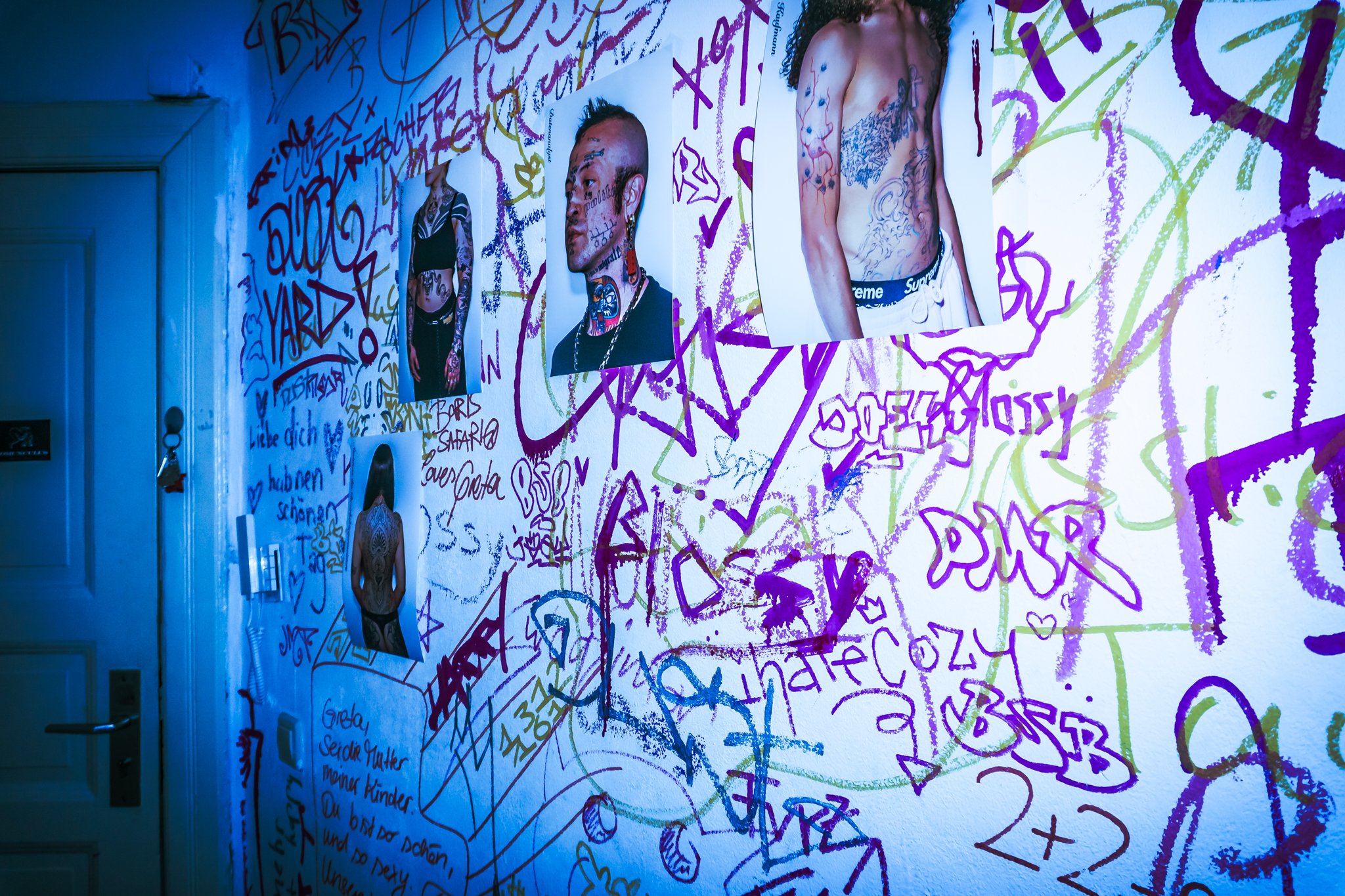 Bild zeigt eine Wand mit bunten Graffiti, Fotos von tätowierten Menschen und handgeschriebene Texte in Deutsch.