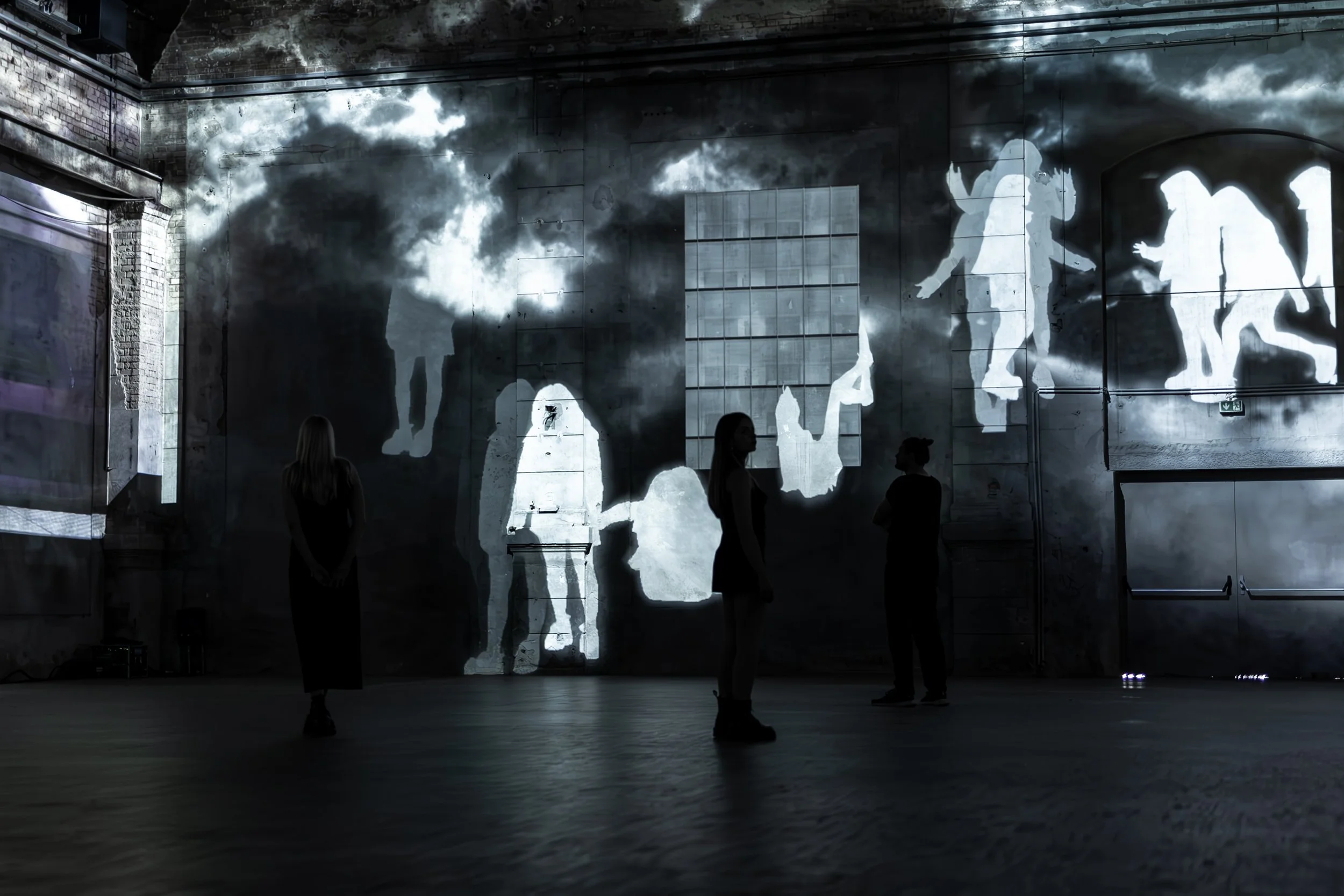 Dunkle Galerie mit Wandprojektion von Silhouetten, drei Menschen stehen vor der Wand, die Silhouetten zeigen Figuren in Bewegung.