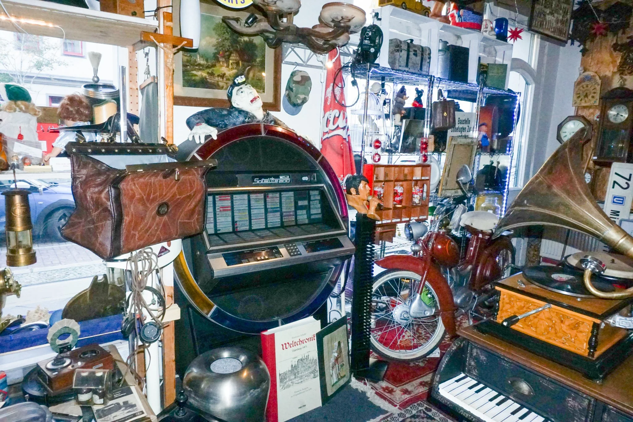 Ein Raum voller verschiedene alte Gegenstände, u.a. eine alte Garderobe, eine Uhr, ein Fahrrad, ein Klavier, Figuren und Dekorationen, die auf einem Flohmarkt oder in einem Antiquitätengeschäft ausgestellt sind.