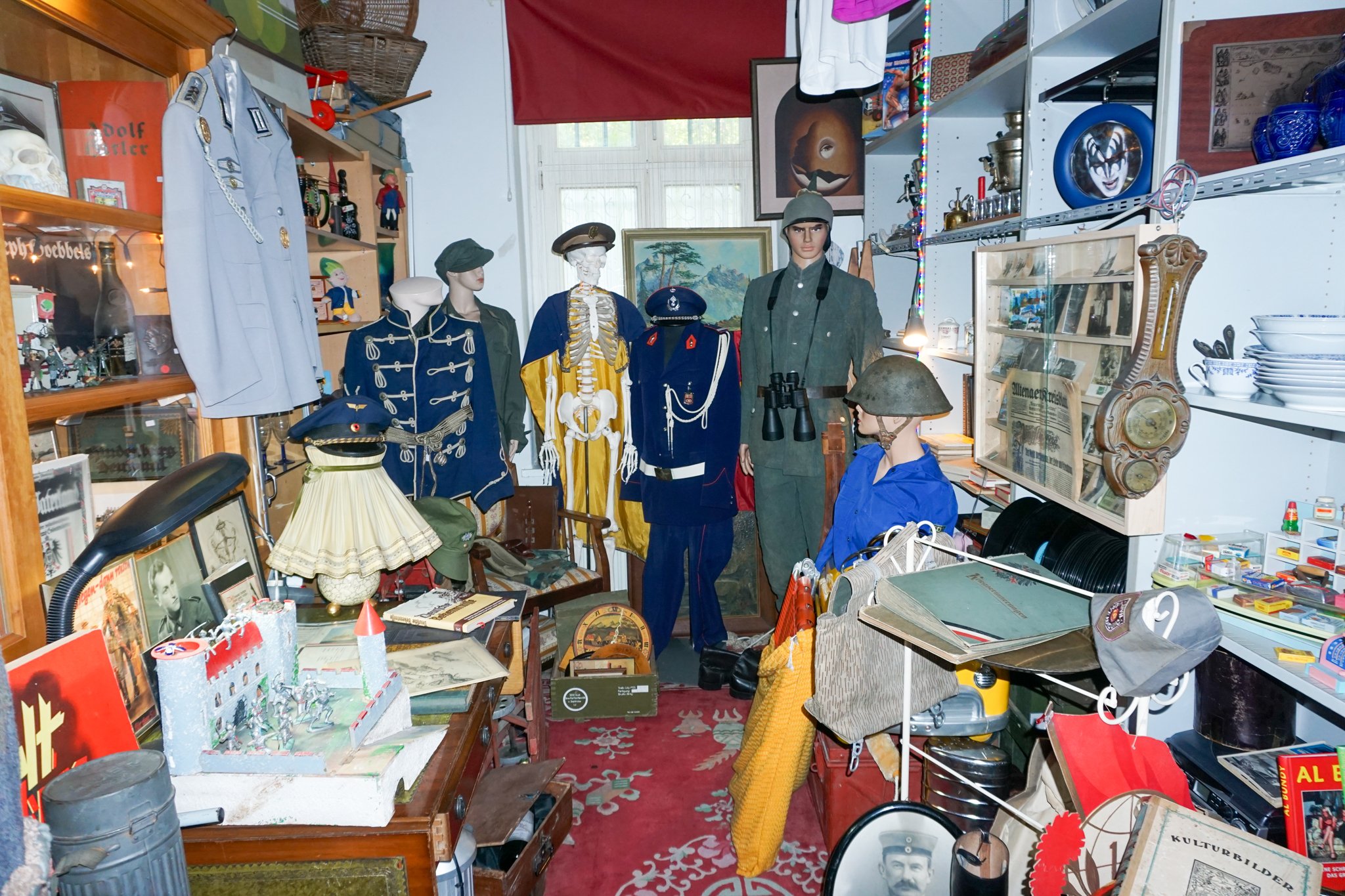 Ein Zimmer mit vielen alten und historischen Gegenständen, darunter mannequin-Körper in Militäruniformen, eine ersatzweise Soldatenfigur, Bilder, Bücher, und verschiedene Dekorationsartikel sowie Spielsachen und Sammlerstücke. Die Ablageflächen sind 