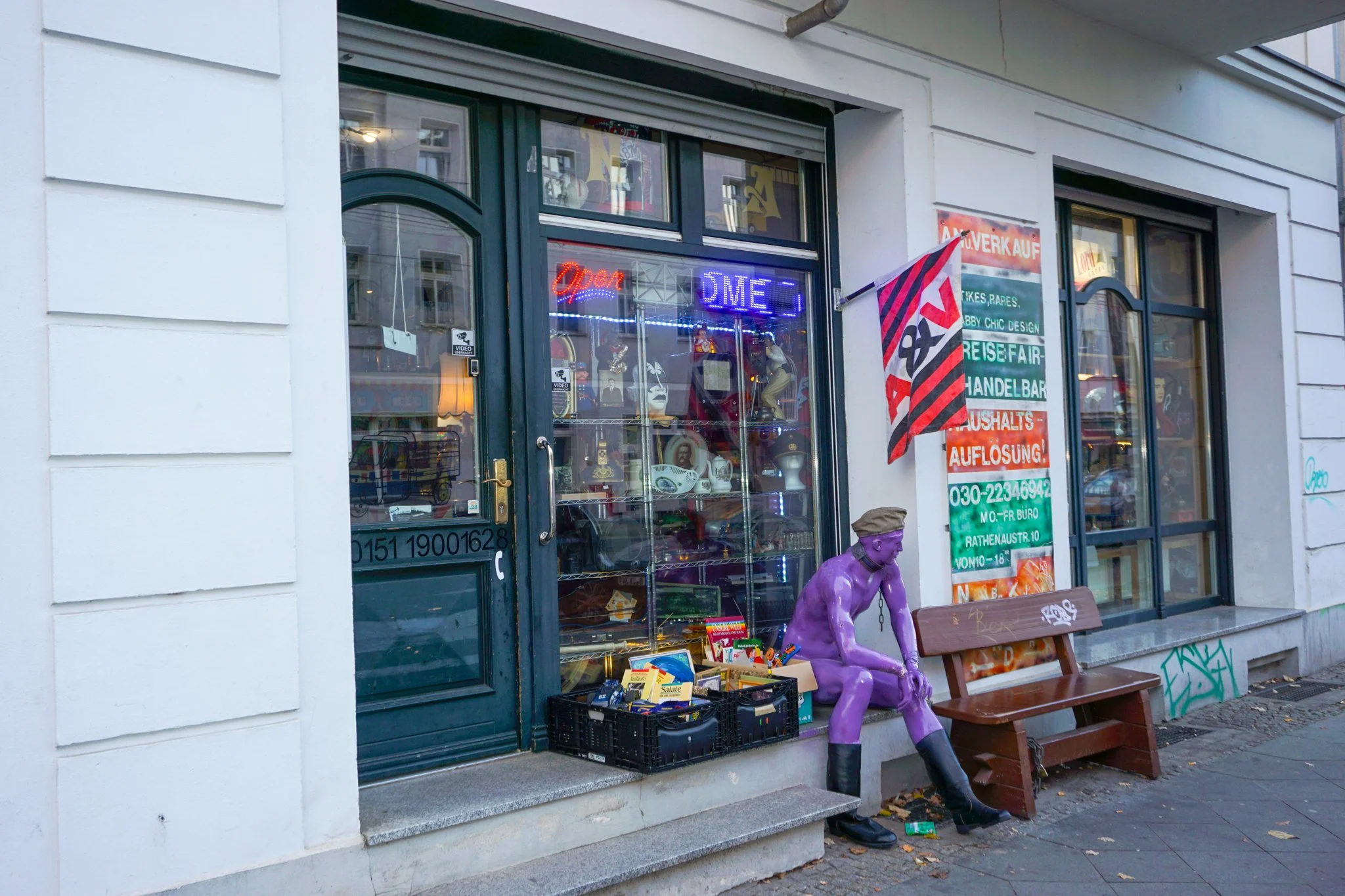 Schaufenster eines Secondhand-Ladens mit Neonbeschriftung 'Open' und 'DME'. Außenszene zeigt einen Sitzbereich mit einer pinkfarbenen sitzenden Statue, einer leeren Bank und einem Poster an der Wand.