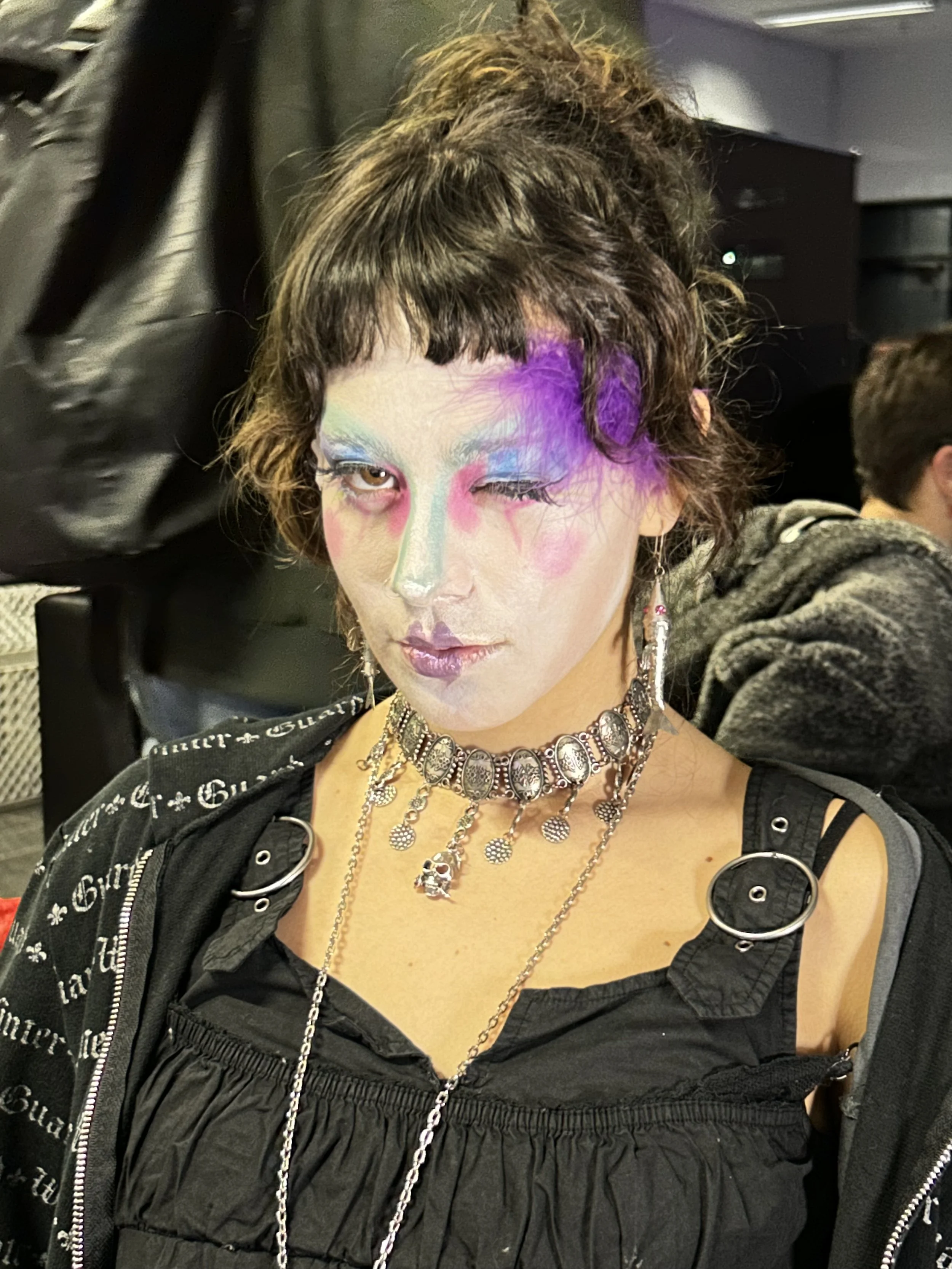 Person mit aufwendigem Make-up und mehreren Ketten, trägt schwarze Kleidung mit Reißverschlüssen, sitzt in einem Raum mit schwarzen Möbeln.