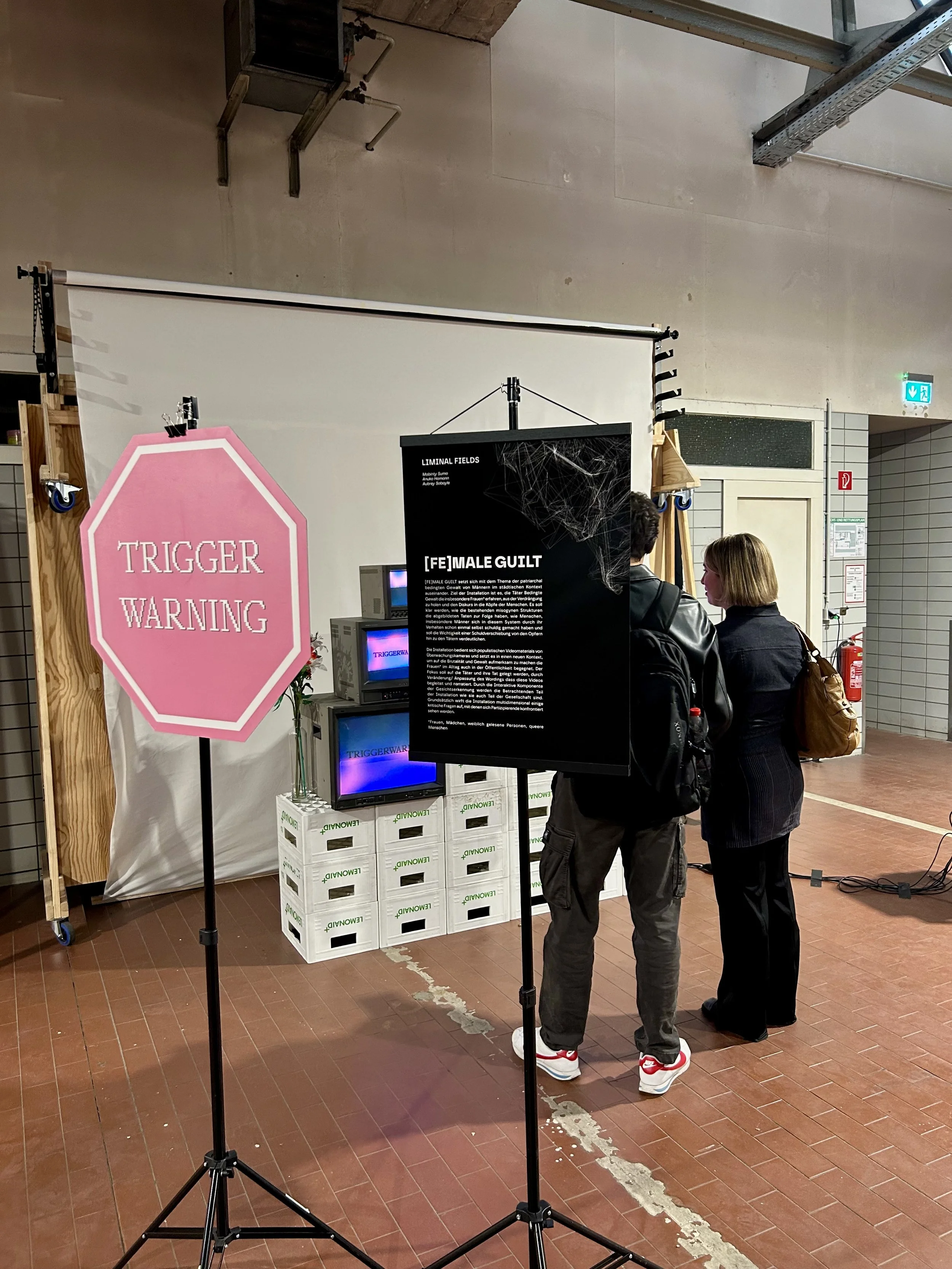 Zwei Menschen stehen vor einem schwarzen Informationstafel im öffentlichen Raum. Es gibt eine große pinkfarbene Stoppschild-Schilder mit der Aufschrift 'TRIGGER WARNING' und einen weißen Hintergrund, sowie eine alte weiße Leiter, eine Fernsehhardware und mehrere Plastikboxen im Vordergrund.