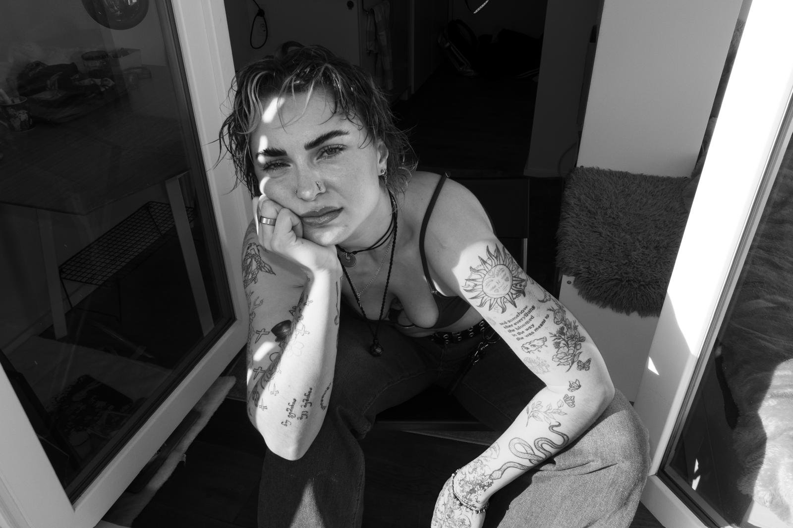Junge Frau mit Tattoos und Piercings sitzt auf einem Stuhl, lehnt den Kopf auf die Hand und schaut in die Kamera, im Hintergrund ein offener Raum mit Rucksack und Kleidung.