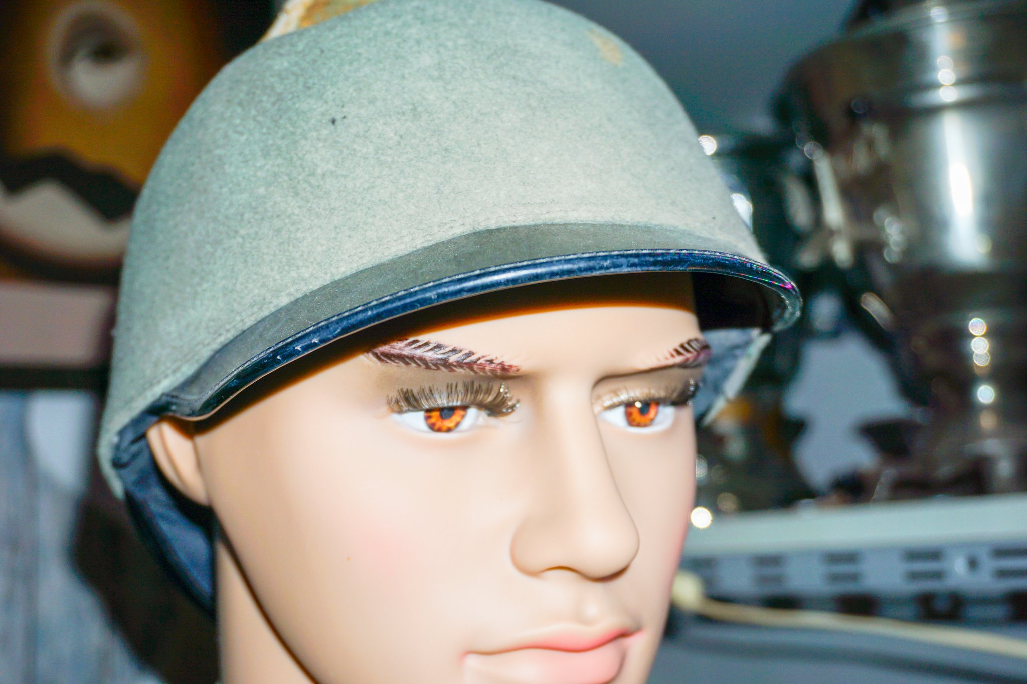Puppe mit braunen Augen, lange Wimpern und blonder Perücke, trägt eine grau-grüne Mütze.