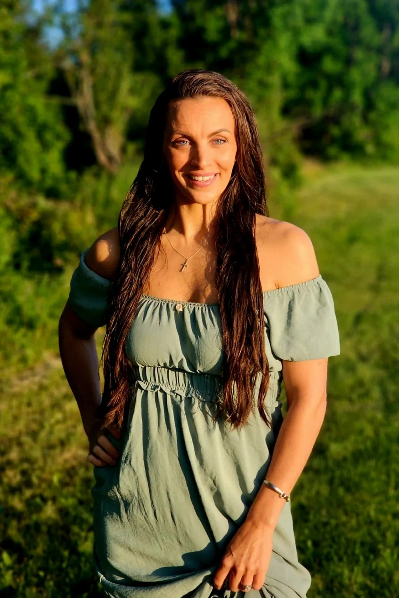 A smiling woman with long, brown hair wearing a light green off-shoulder dress, standing outdoors in a sunny, green field.