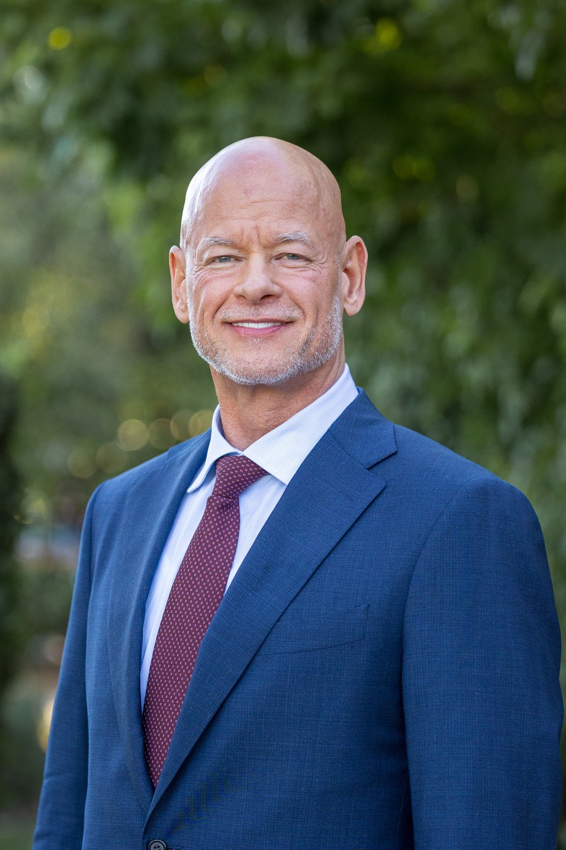 A smiling man with wearing a blue suit, white shirt, and maroon tie, standing outdoors with greenery in the background.