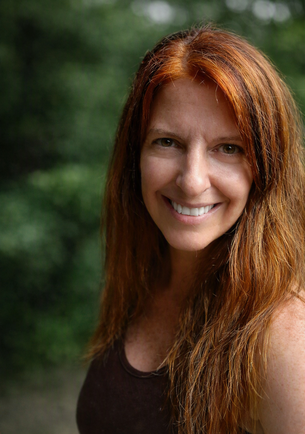 A smiling woman with red hair outdoors with green foliage in the background.