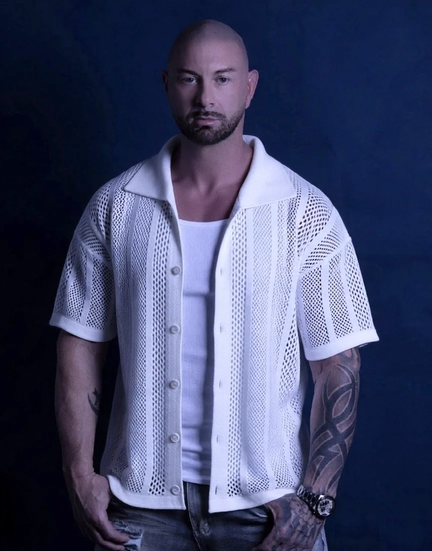 A man with a shaved head and beard posing against a dark background, wearing a white mesh short-sleeve button-up shirt over a white tank top, with tattoos on his forearms, a watch on his wrist, and casual jeans.