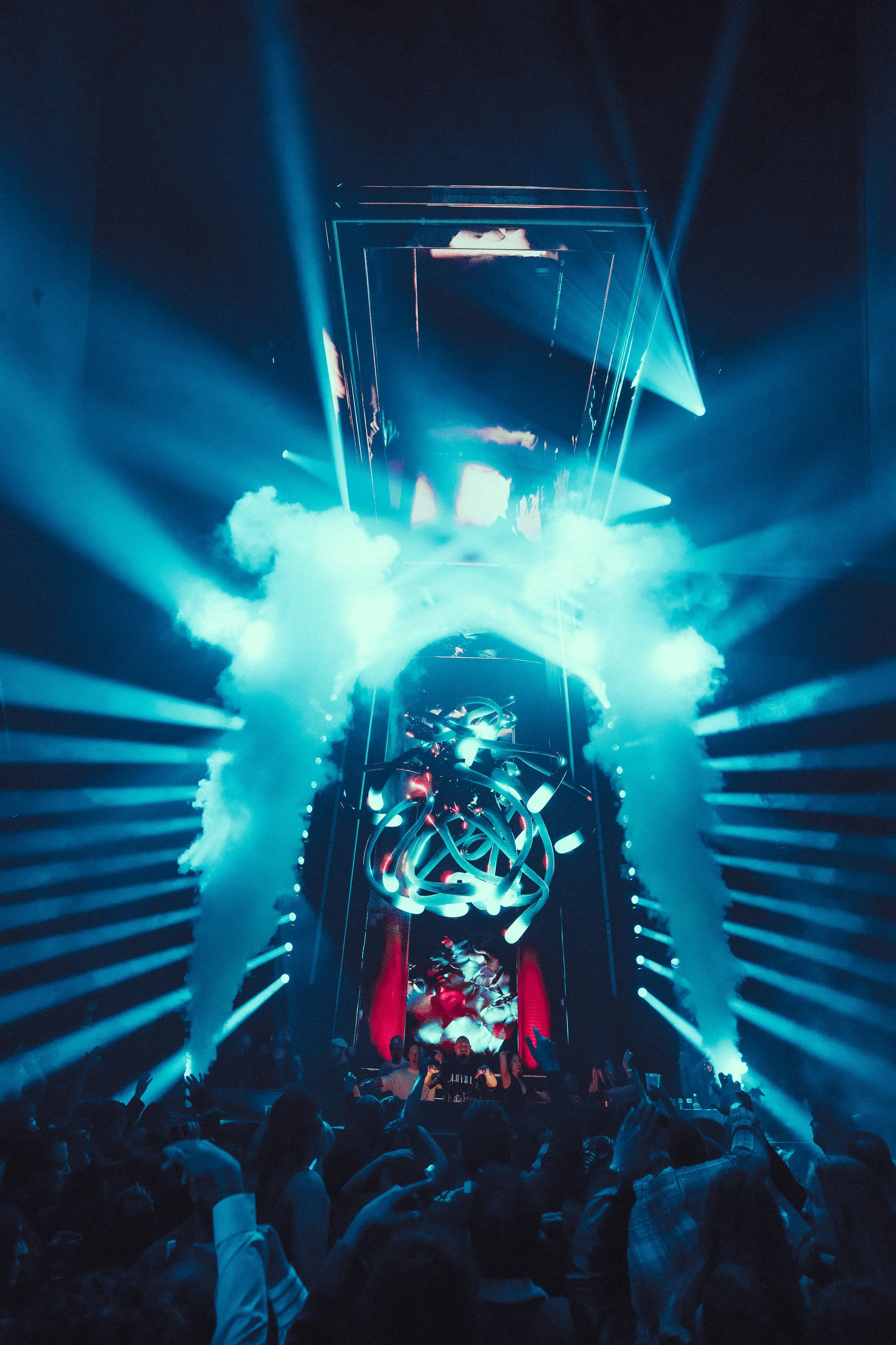 A crowded nightclub scene with blue lighting, smoke effects, and a DJ performing on stage surrounded by neon lights and visual projections. Many people are dancing and taking photos.