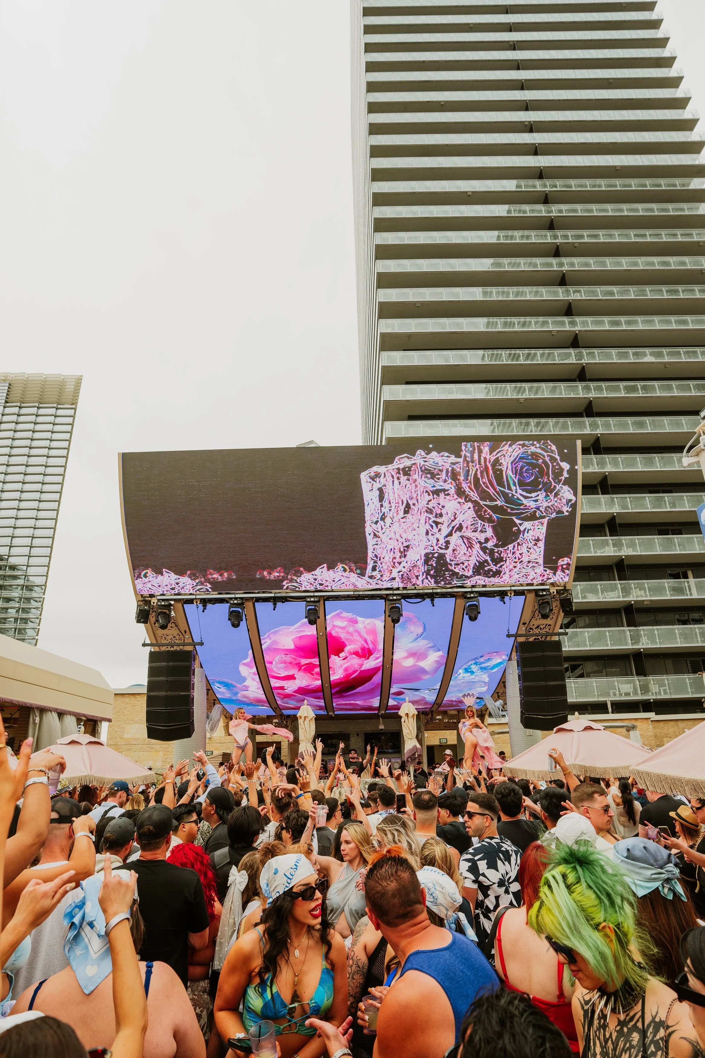 Crowd of people at outdoor event with large digital screens showing colorful floral visuals, high-rise buildings in the background on cloudy day