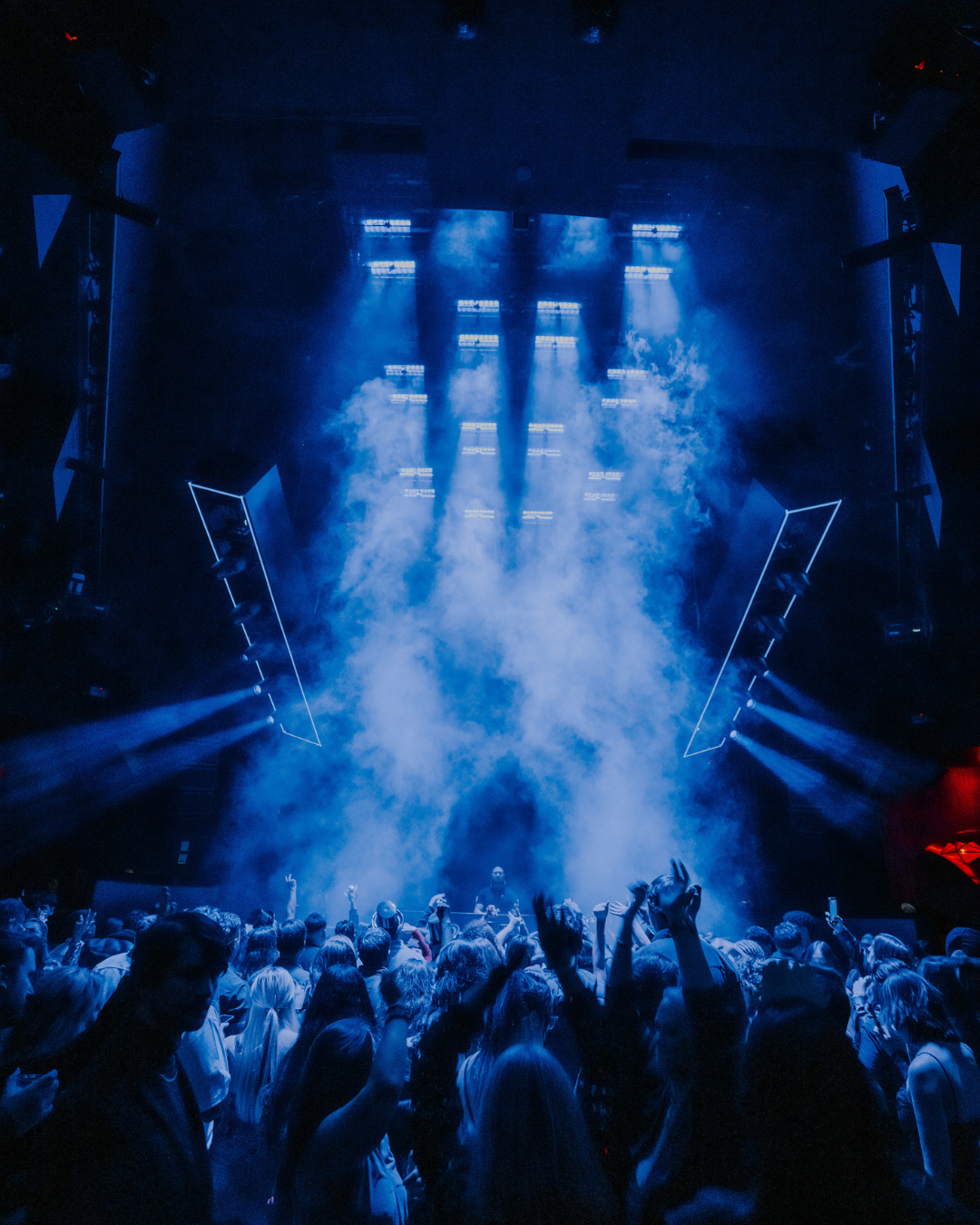 Crowd at a concert or nightclub with blue stage lighting, fog, and DJ or performer on stage.