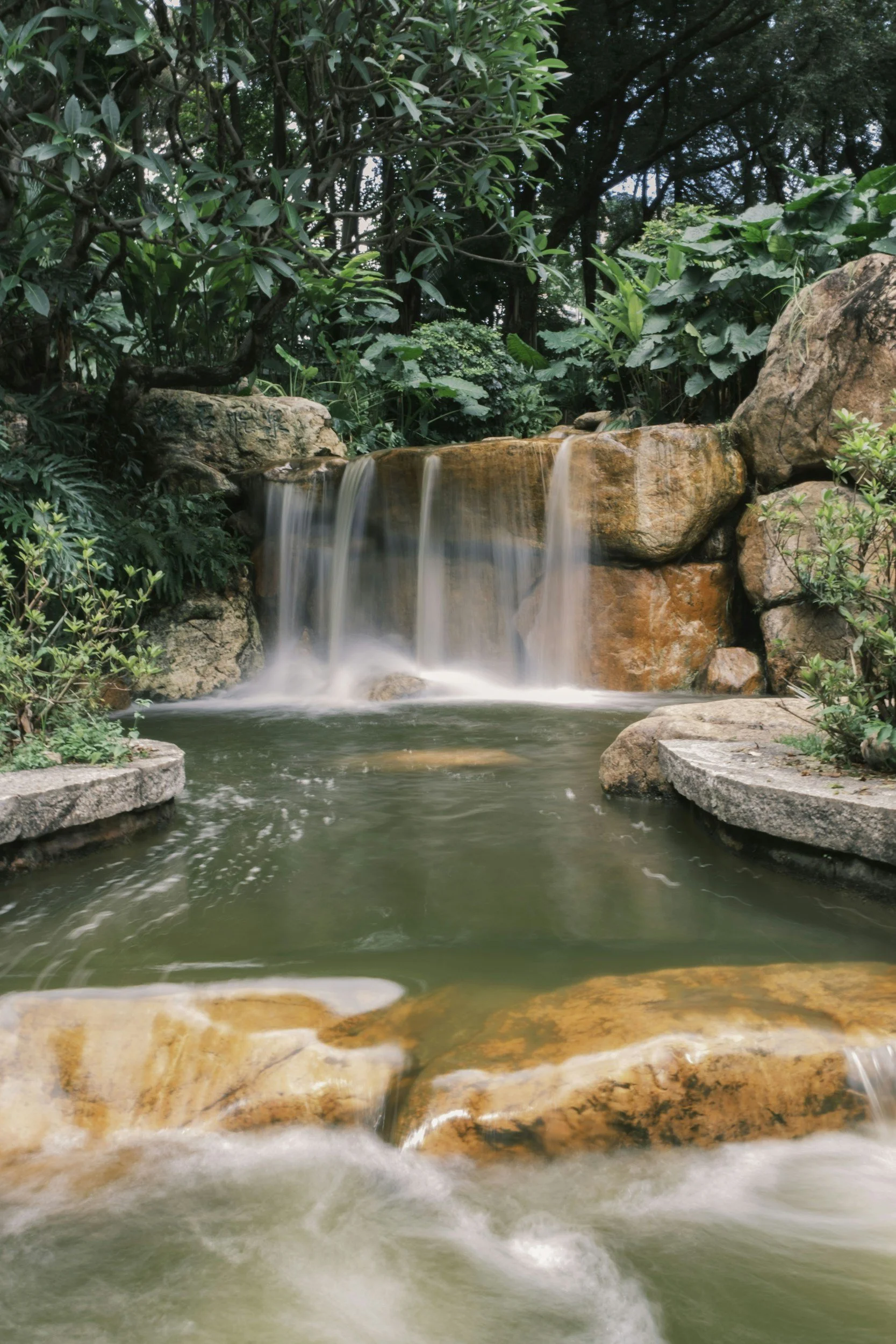 A small waterfall cascading over rocks into a pond, surrounded by lush green plants and trees.