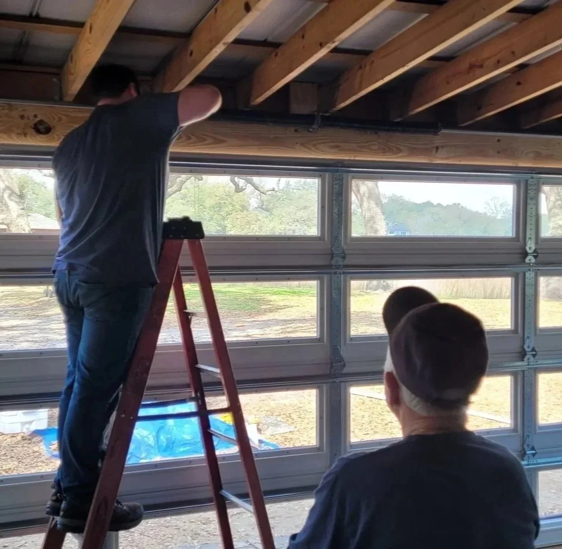 Two people are working on a garage door installation or repair. One person, standing on a ladder, is working on the ceiling, while the other person is watching.