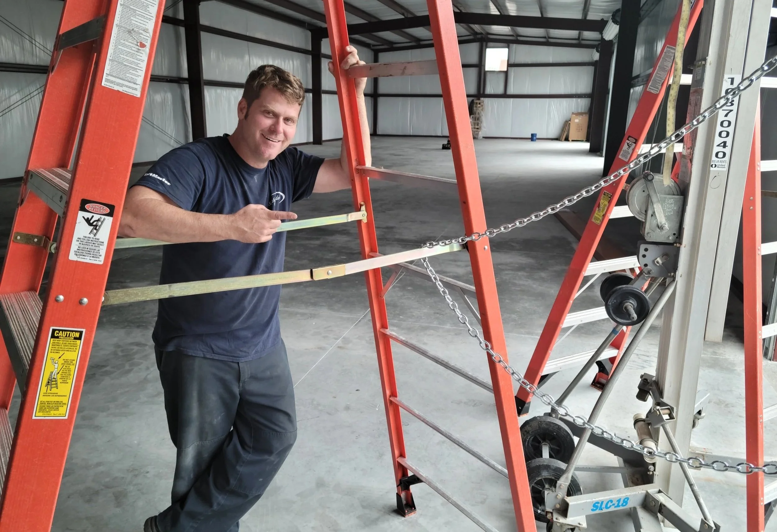 A man smiling and pointing at a safety harness attached to an orange ladder inside a warehouse or industrial building.