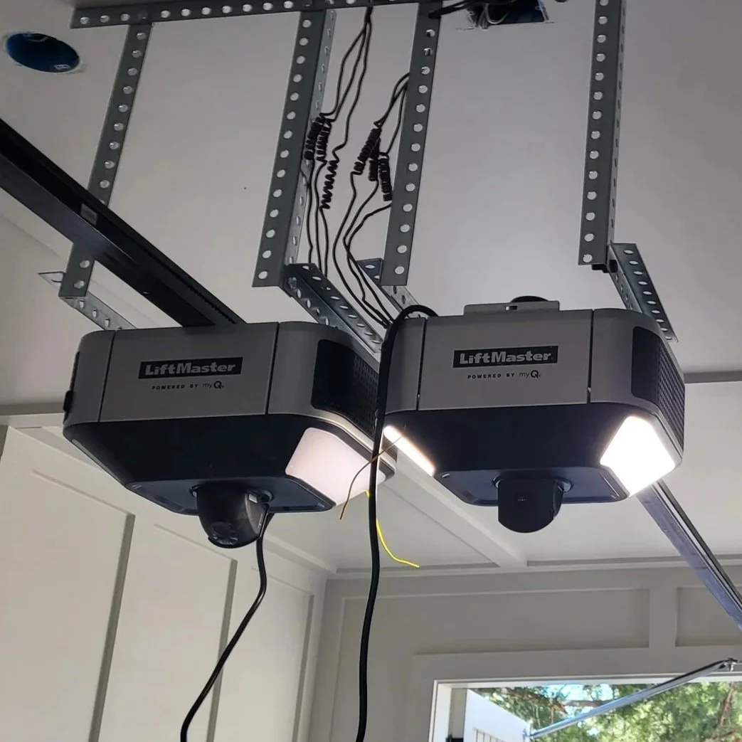 Two LiftMaster garage door openers mounted to a metal track system on the ceiling, with wires hanging down, in a residential garage.