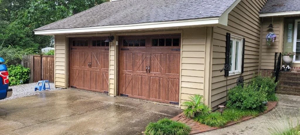 A two-car garage attached to a beige house with a garden bed beside the stairs. The garage doors are wooden with black handles and windows at the top. There's a small light above the garage doors.