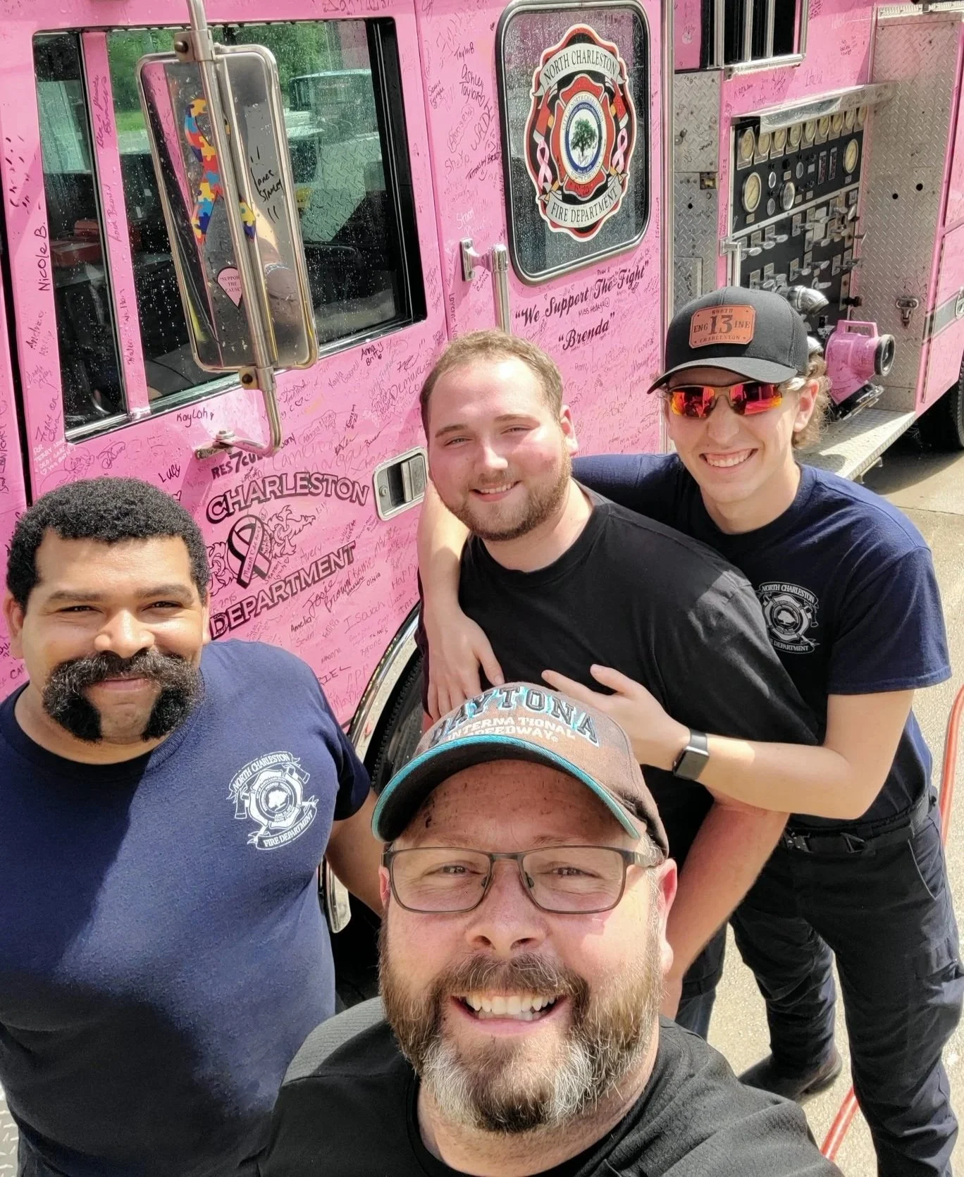 Group of five men taking a selfie in front of a pink fire truck with signatures and messages. The men are smiling, and the fire truck features the North Charleston Fire Department emblem.