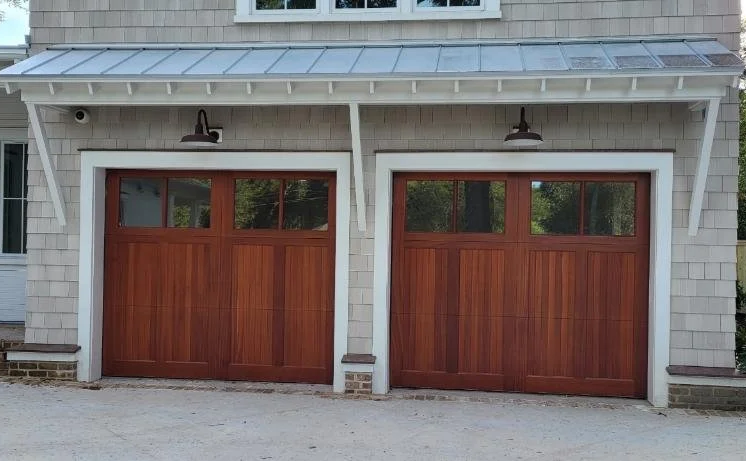 Two wooden garage doors with small windows at the top, framed by white trim, under a metal awning, part of a house with light-colored siding.