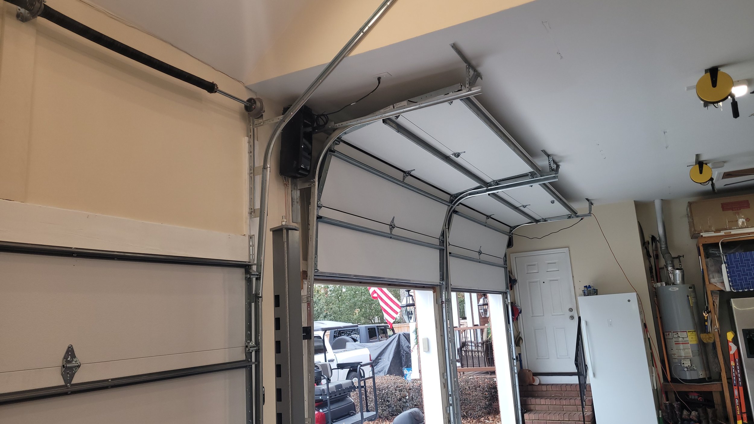 Inside a home garage with an open garage door, showing a high lift garage door opener system and some tools and boxes on shelves.