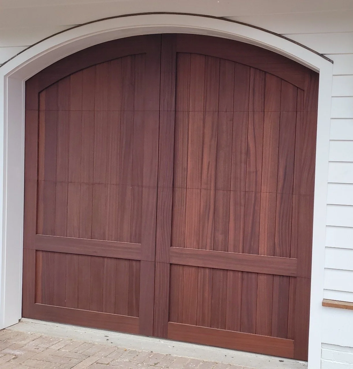 A large double wooden garage door with an arched top, set in a white building with siding, on a brick-paved area.