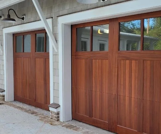 Two wooden garage doors with small windows at the top, set in a house with white framing and gray exterior siding.