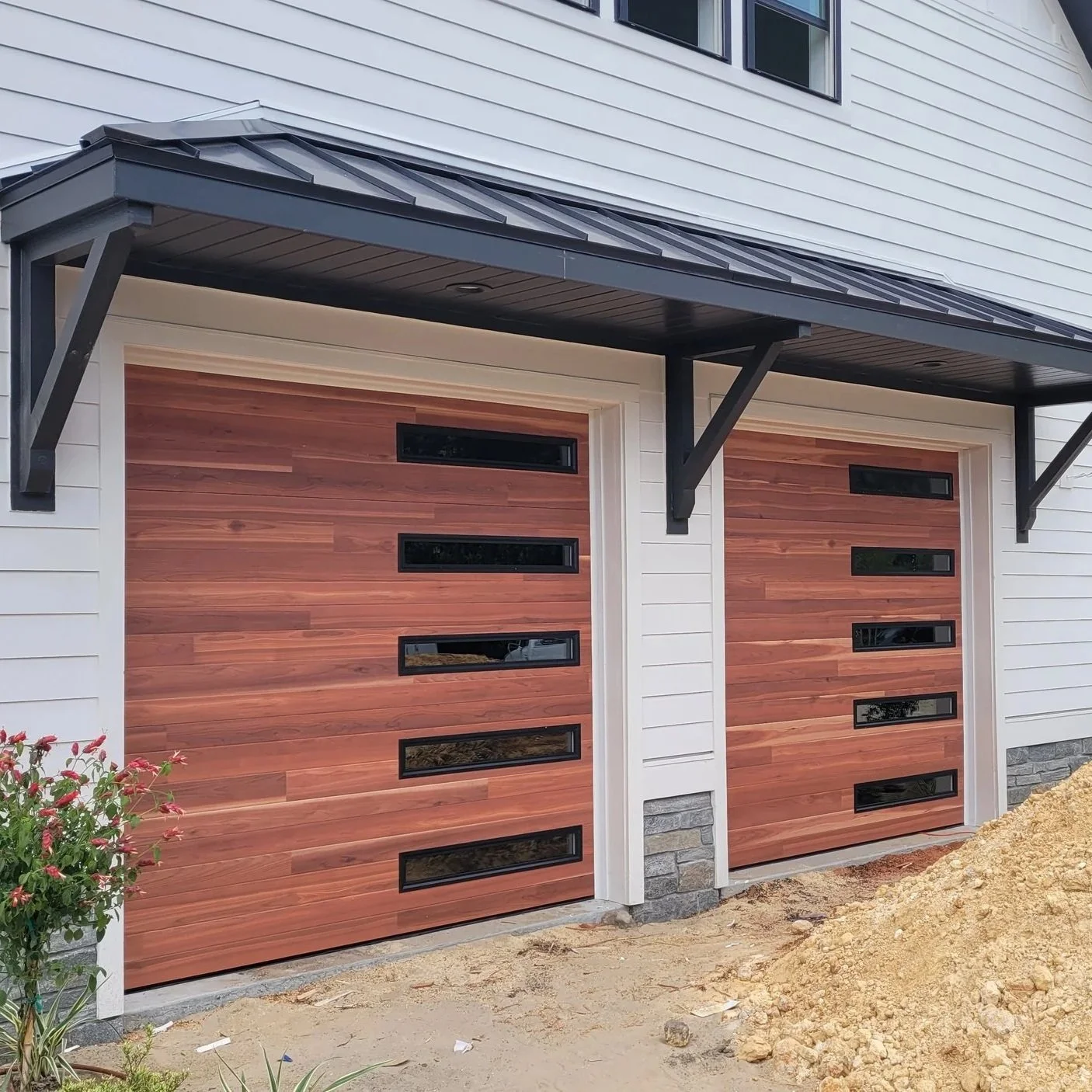 Two wooden garage doors with horizontal glass inserts under a black metal awning on the exterior of a house with white siding and small windows.