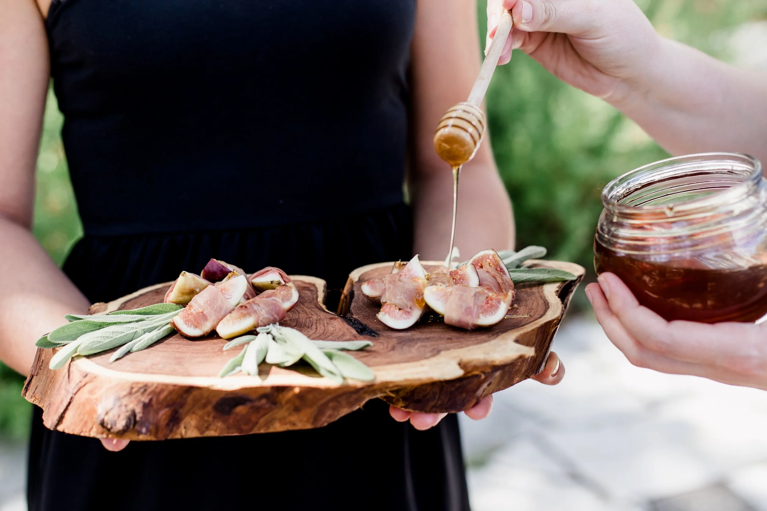 Person holding a wooden serving board with sliced figs, fresh herbs, and honey being drizzled on top, with another person pouring honey from a jar.