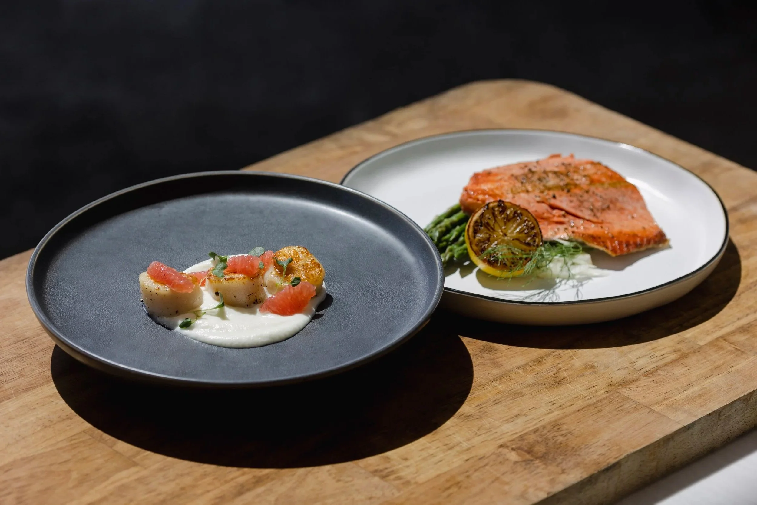 Two plates of plated food on a wooden board: one with seared scallops garnished with microgreens and pink grapefruit segments, the other with a piece of grilled salmon served with asparagus, a halved grilled lemon, and fresh herbs.