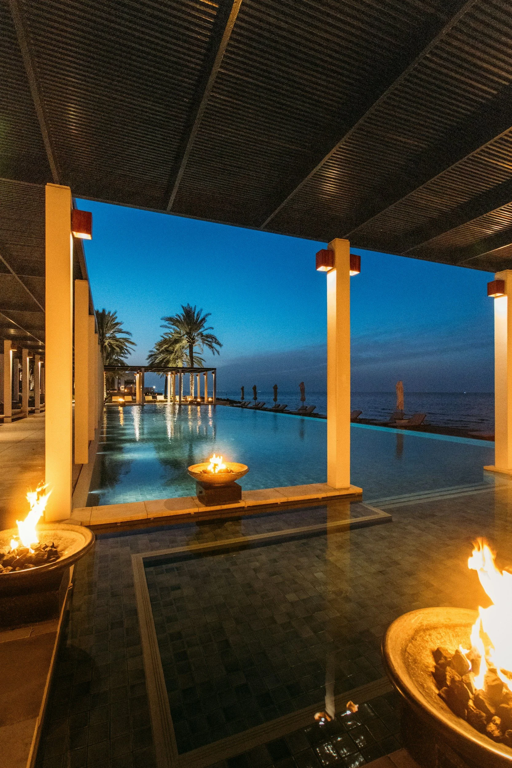 An outdoor pool area at dusk with palm trees in the background, lit fire pits, and modern architecture with columns and a dark ceiling.