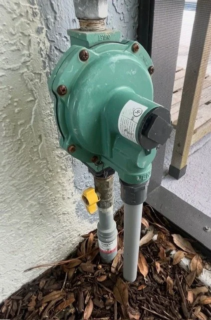 2nd Stage Gas Regulator, installed on a pipe next to a textured wall and a balcony railing, surrounded by dry leaves on the ground.