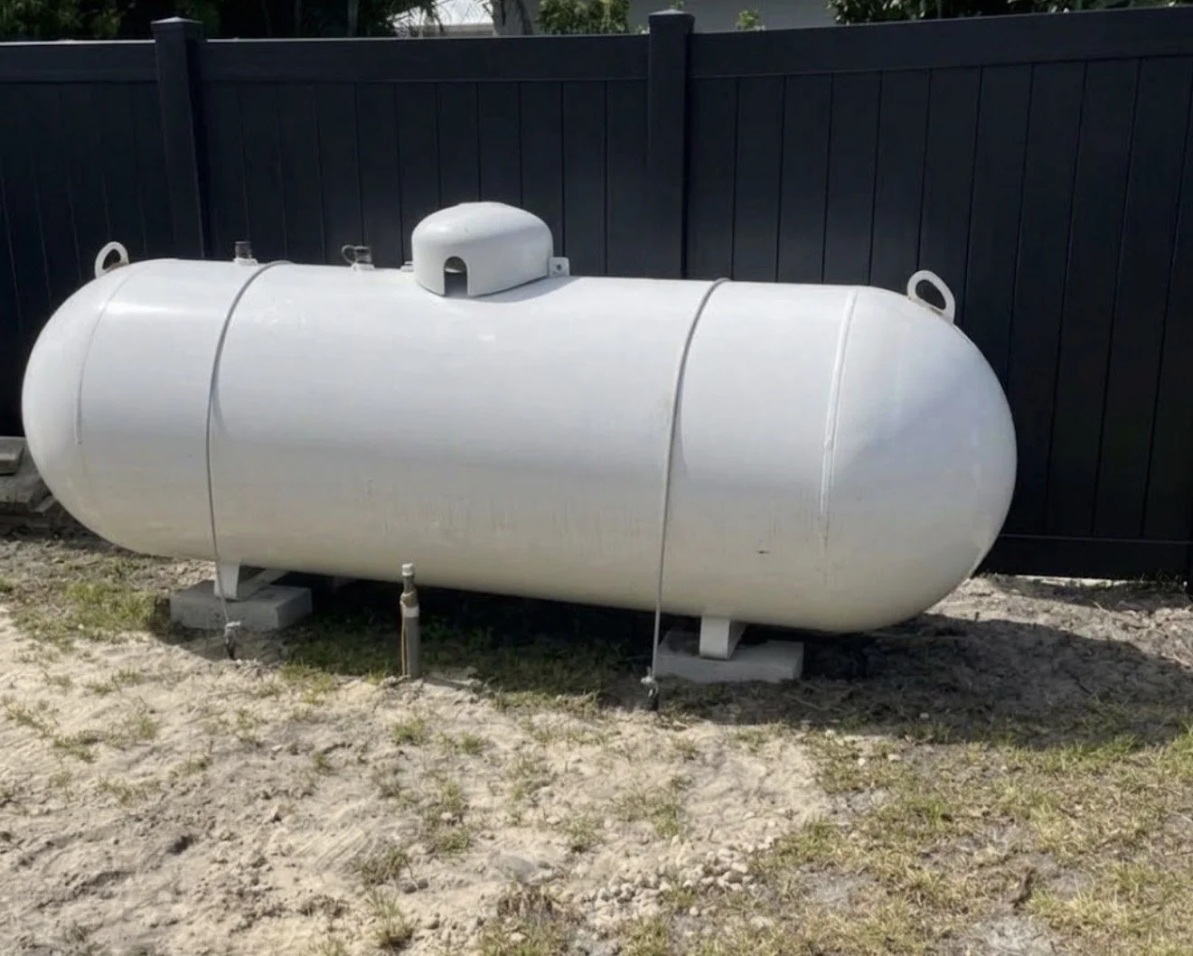 White horizontal propane tank on concrete blocks in a yard with grass and dirt, black fence in the background.