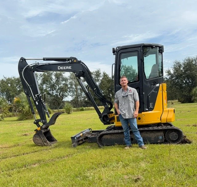 A man standing next to a small black and yellow John Deere excavator on a grassy field with trees in the background.