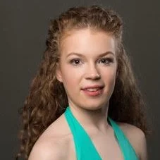 Portrait of a young woman with curly hair wearing a turquoise top, smiling softly against a gray background.