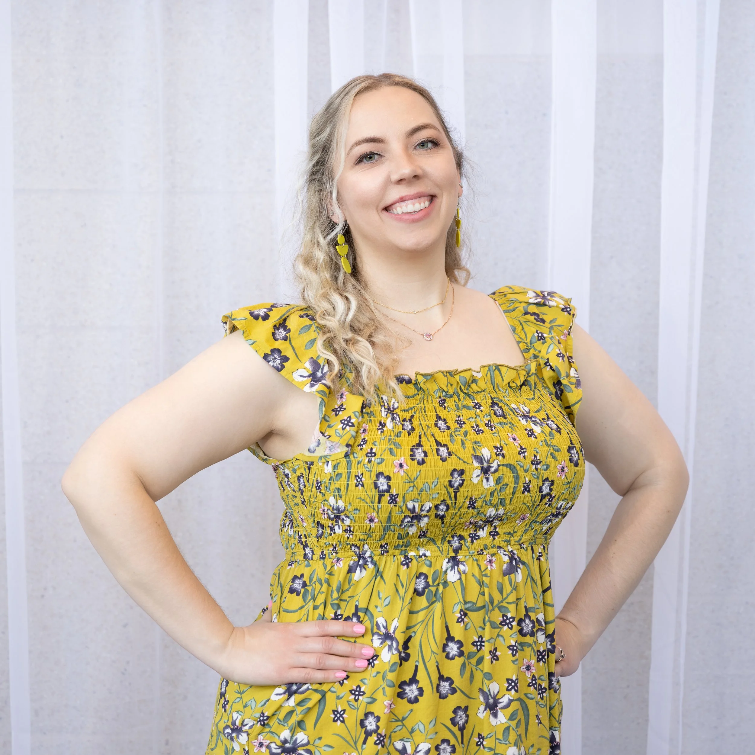 Close-up of a young woman with long, wavy blonde hair smiling and wearing yellow floral dress with ruffled sleeves, standing against a light-colored wall.