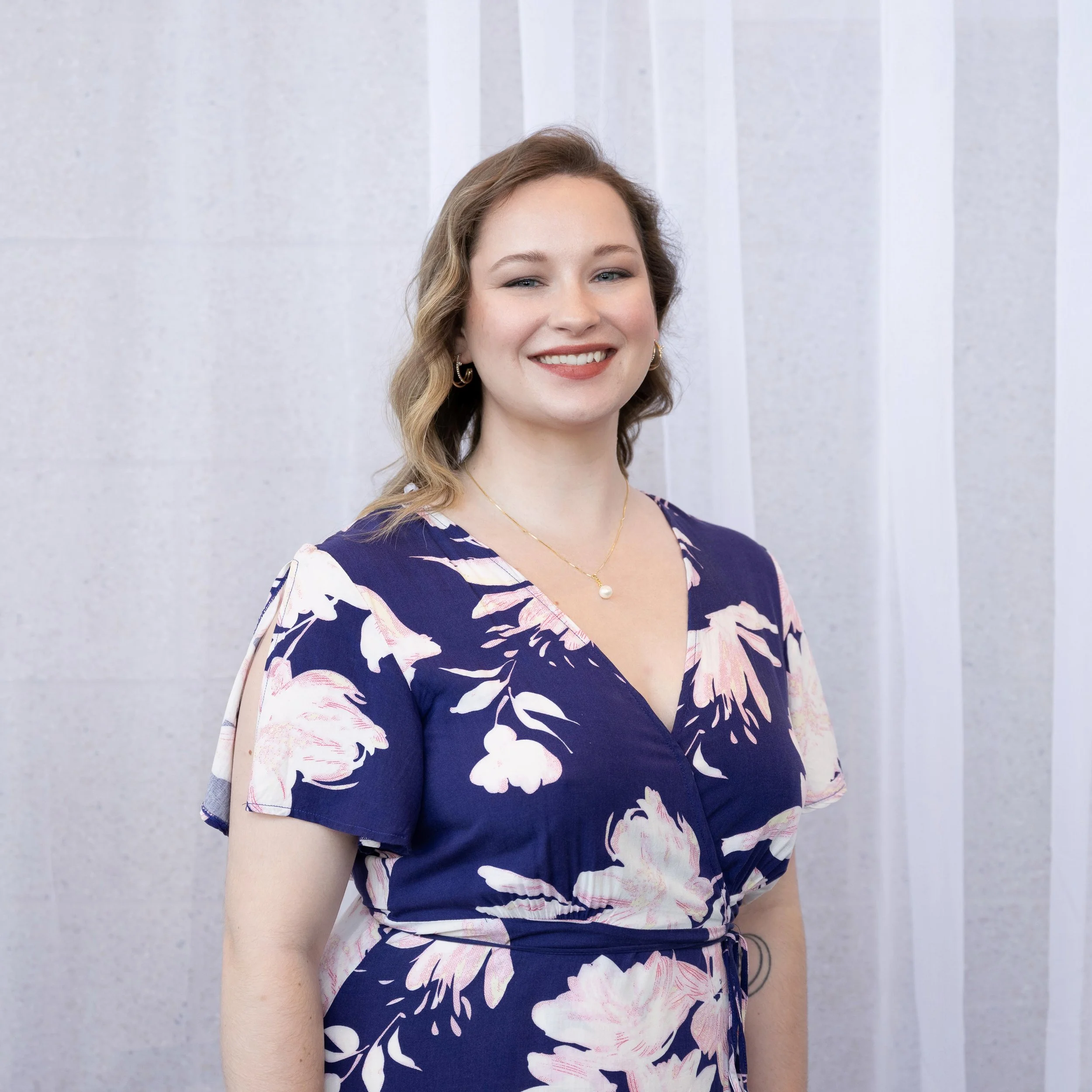 A young woman with wavy light brown hair, wearing a navy blue floral dress with pink and white flowers, gold hoop earrings, and a pearl necklace, smiling in front of a white, slightly textured background.