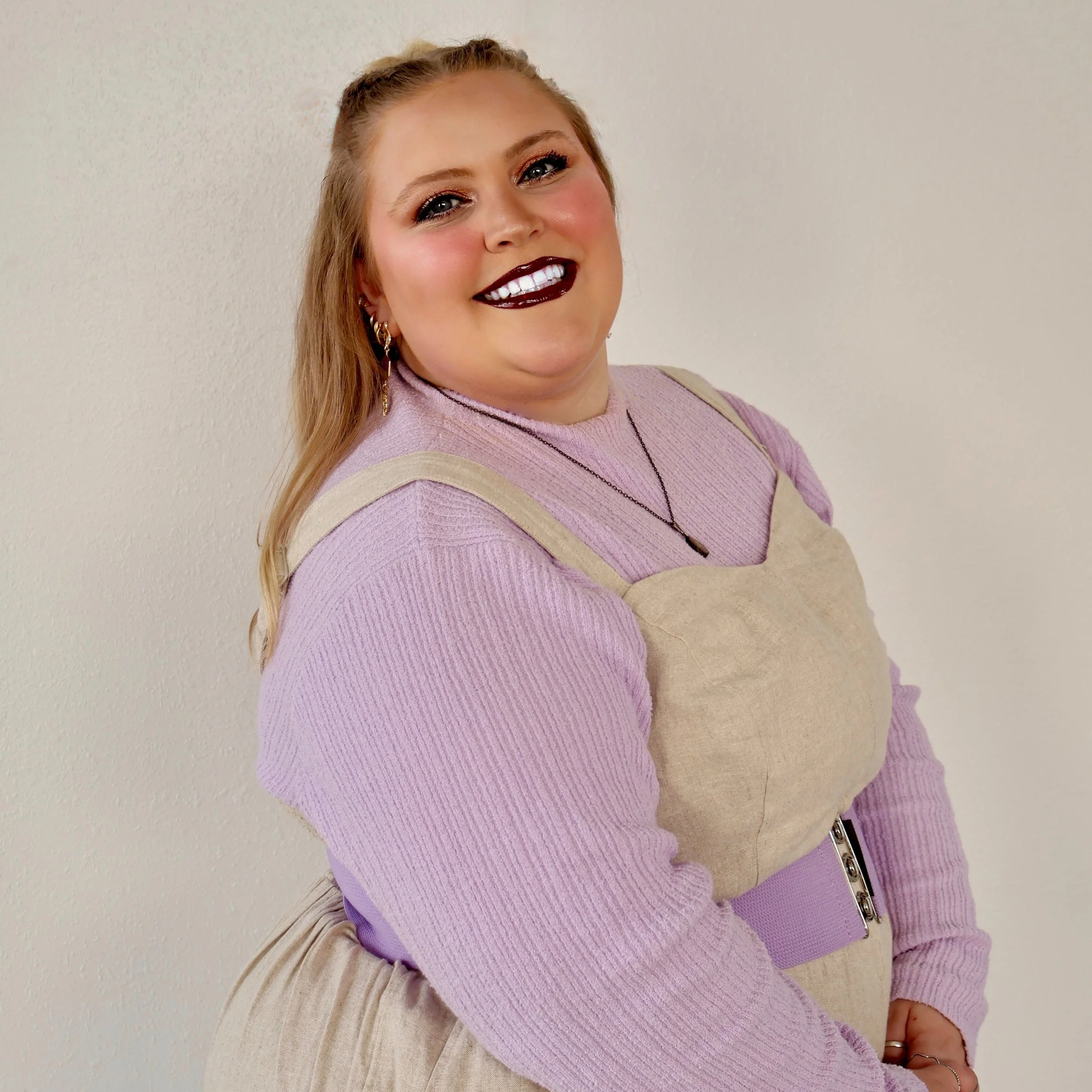 A young woman with long blonde hair styled in a ponytail is smiling at the camera. She has bright blue eyes, dark makeup, including dark lipstick, and is wearing gold earrings, a necklace, a light purple sweater, and beige overalls. She is standing against a plain light-colored wall.