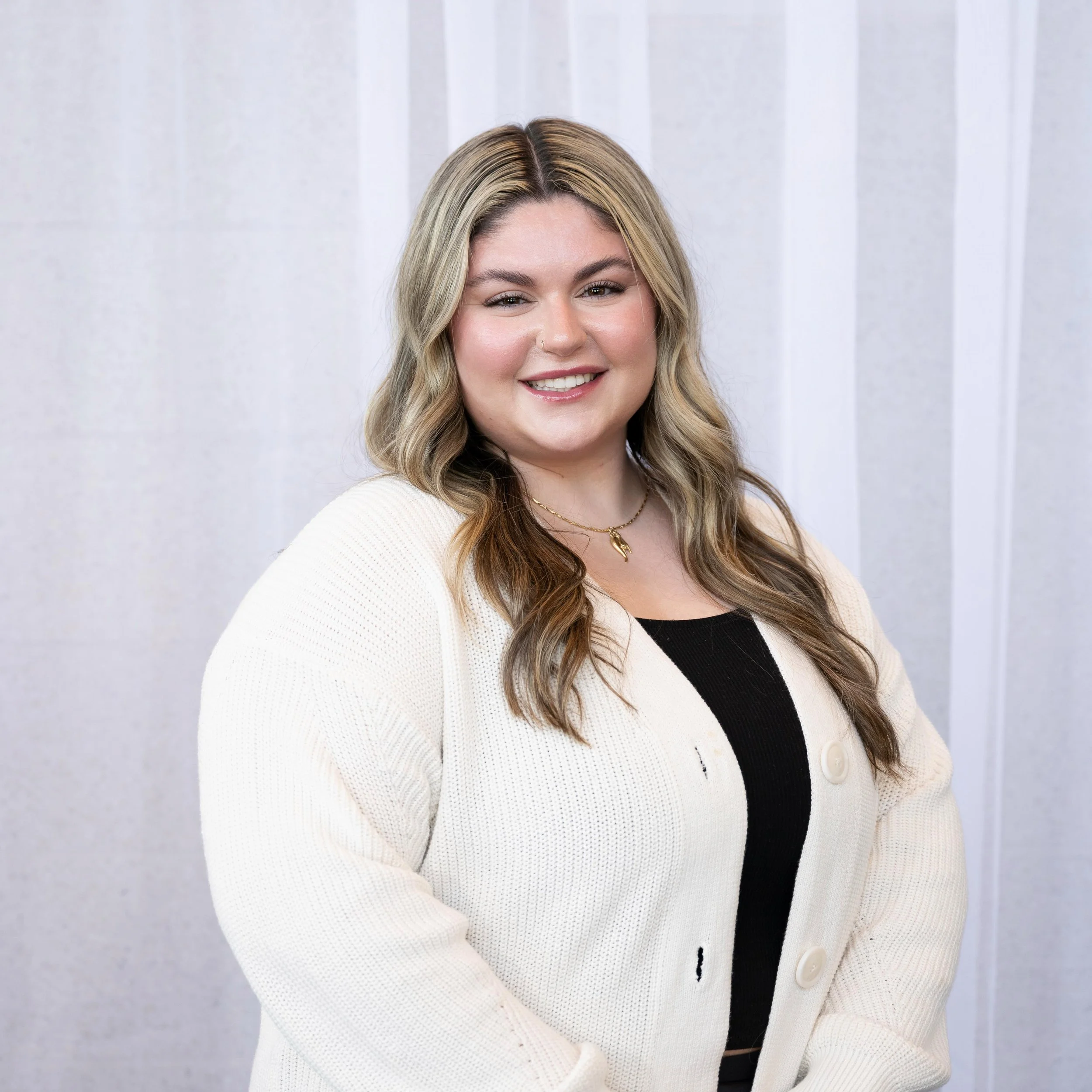 A smiling woman with long, wavy blonde hair wearing a white knit cardigan over a black top, standing in front of a light-colored striped background.